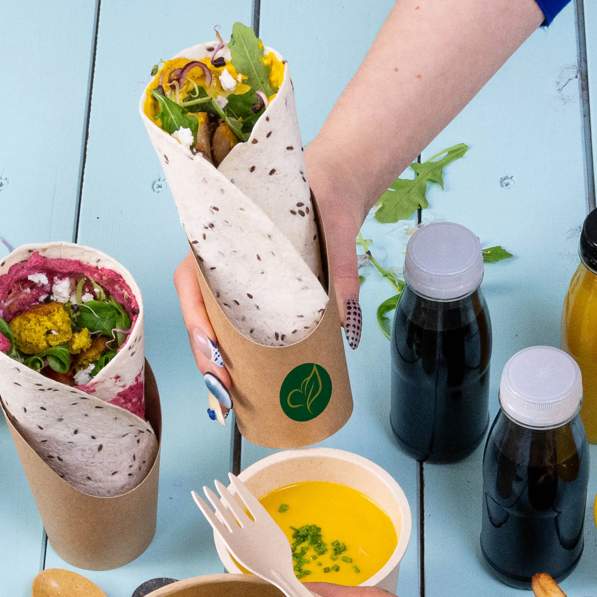 Un gobelet en carton kraft contenant un wrap rempli; un autre gobelet avec un wrap coloré à côté, entouré de boissons et d'une sauce à tremper.