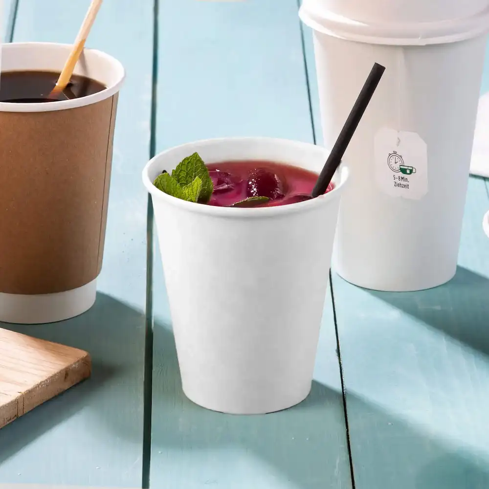 Gobelet en papier blanc contenant une boisson avec des feuilles de menthe, une paille noire et deux autres gobelets en arrière-plan.