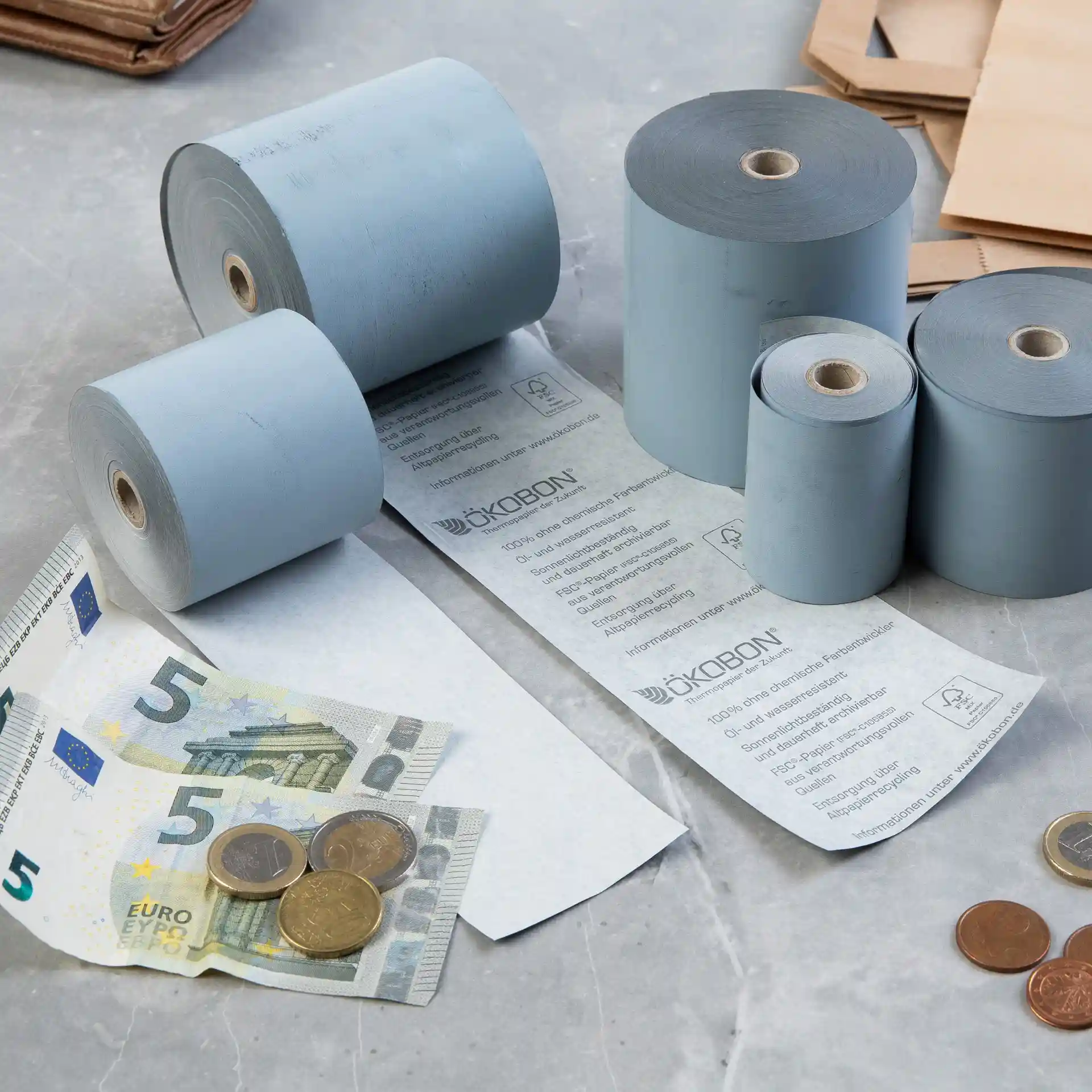 Image de plusieurs rouleaux de papier thermique gris, d'un billet de 5 euros et de pièces sur une surface.