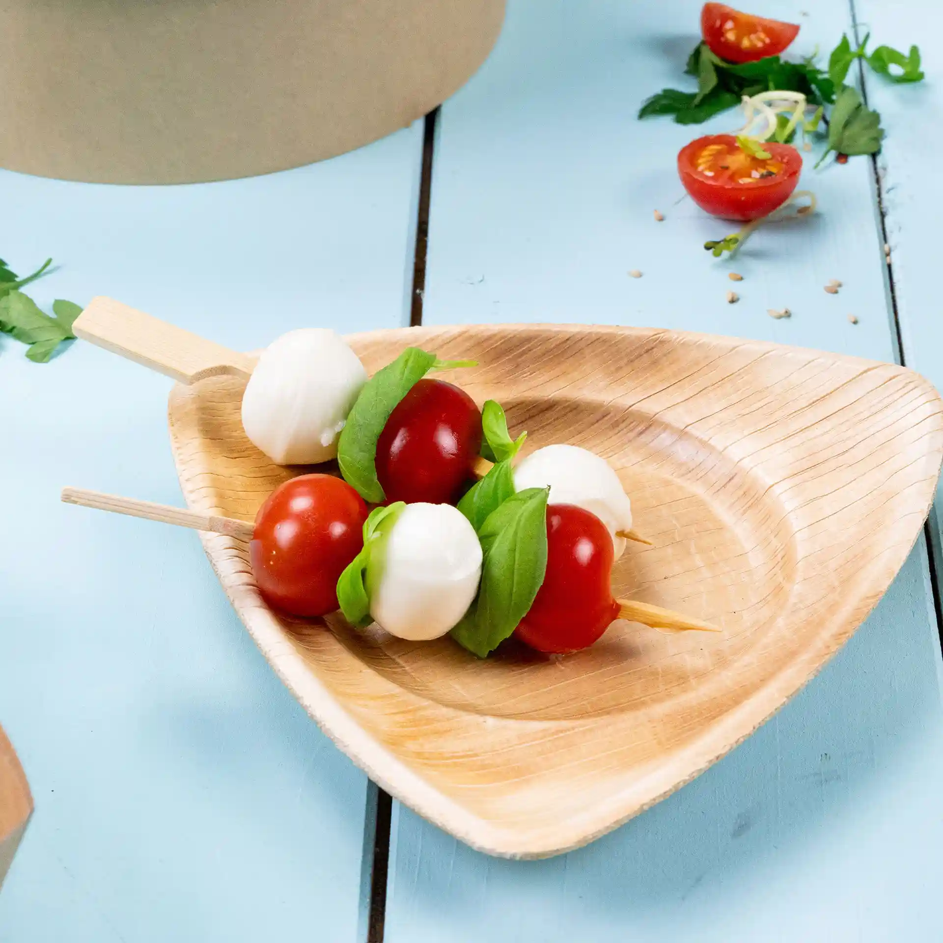 Brochettes en bambou avec tomates cerises, billes de mozzarella et basilic sur une assiette en bois triangulaire.