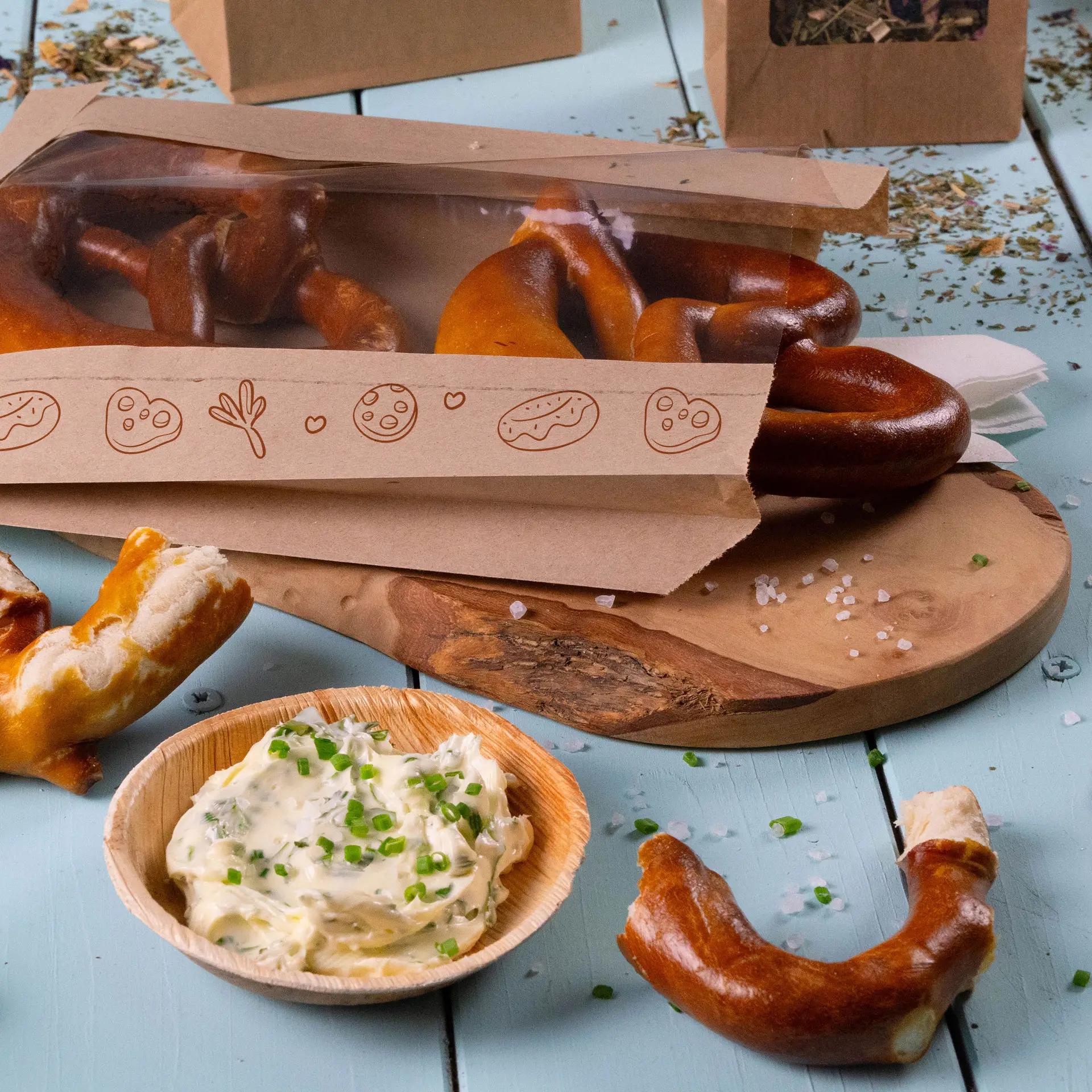 Un sac en papier imprimé avec fenêtre contenant des bretzels moelleux ; petit plat de dip crémeux aux herbes vertes à côté sur une planche en bois.