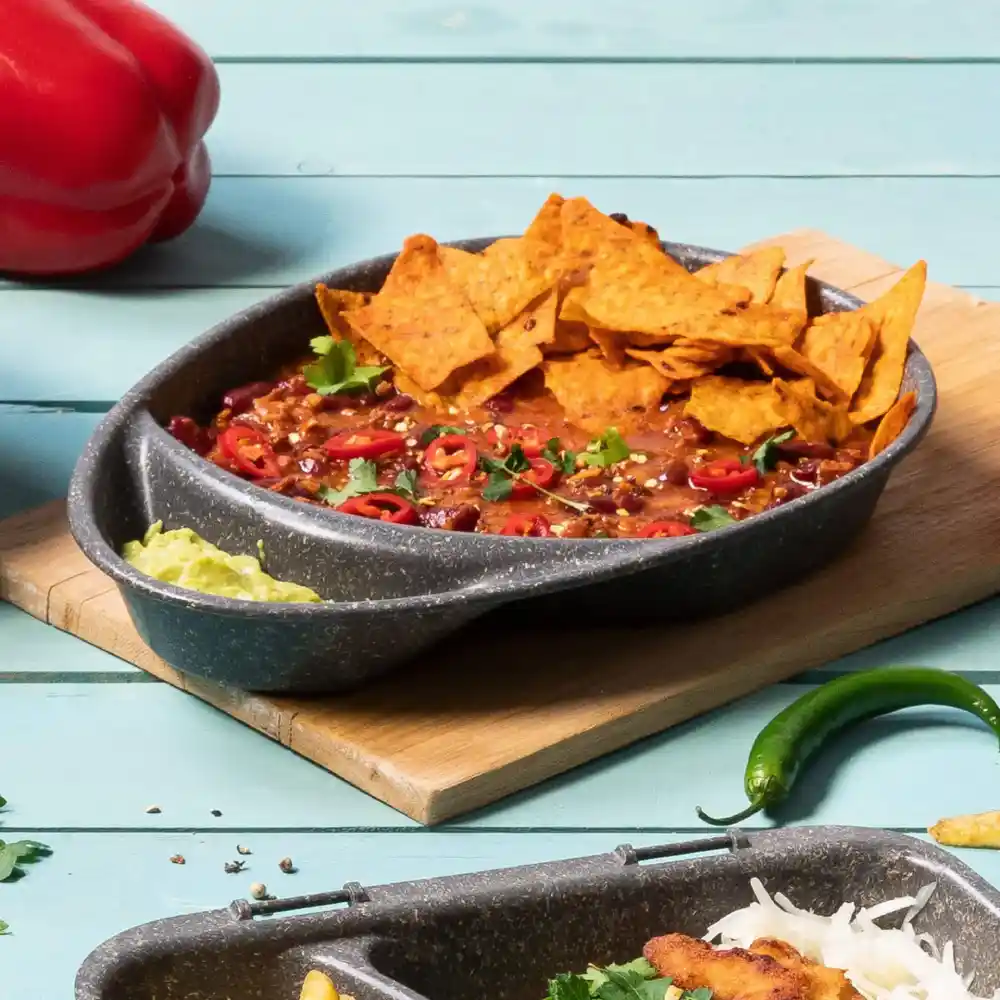 Bol de chili garni de chips tortilla, piments rouges tranchés et coriandre sur une planche en bois avec garnitures.