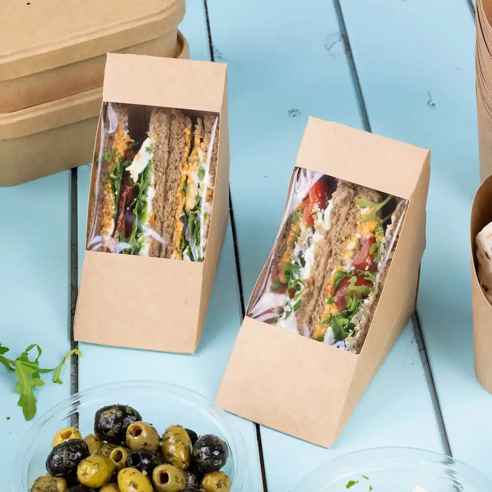 Deux boîtes à sandwich en papier kraft brun avec des fenêtres montrant des sandwiches en couches et un bol d'olives mélangées.