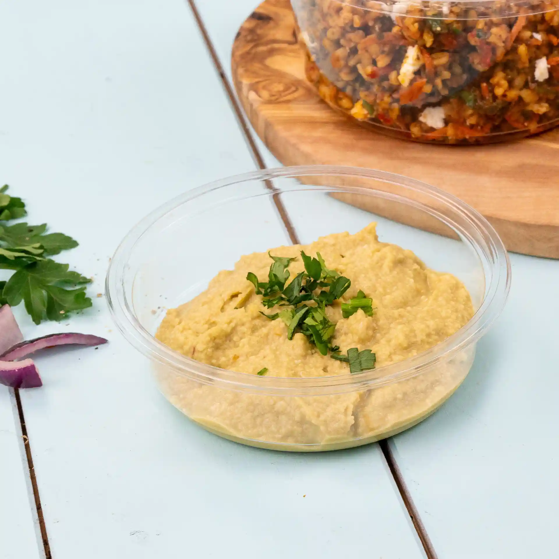 Conteneur deli transparent rond avec du houmous garni de persil haché, placé sur une surface bleu clair.