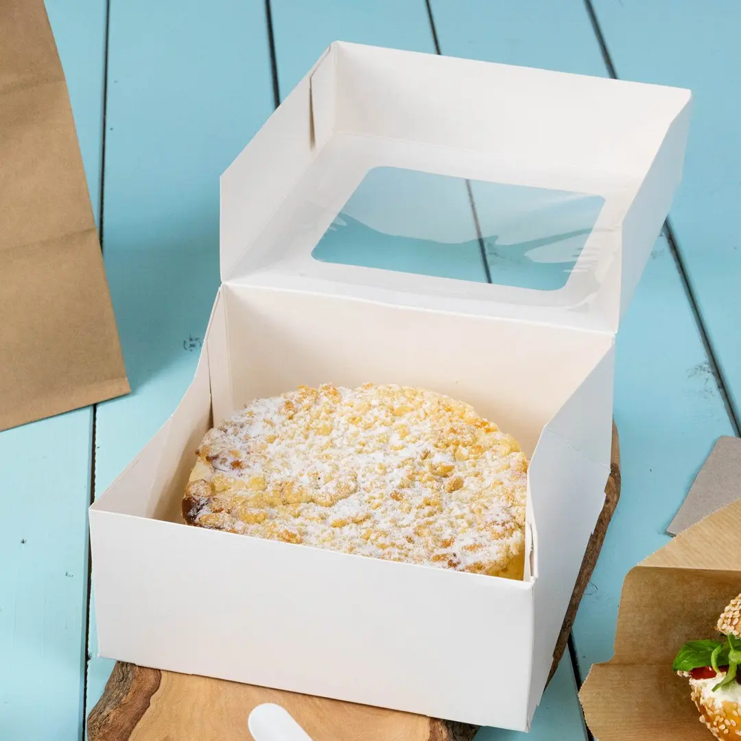 Boîte à gâteau blanche avec une fenêtre transparente, contenant un gâteau rond saupoudré de sucre glace, sur une surface en bois.