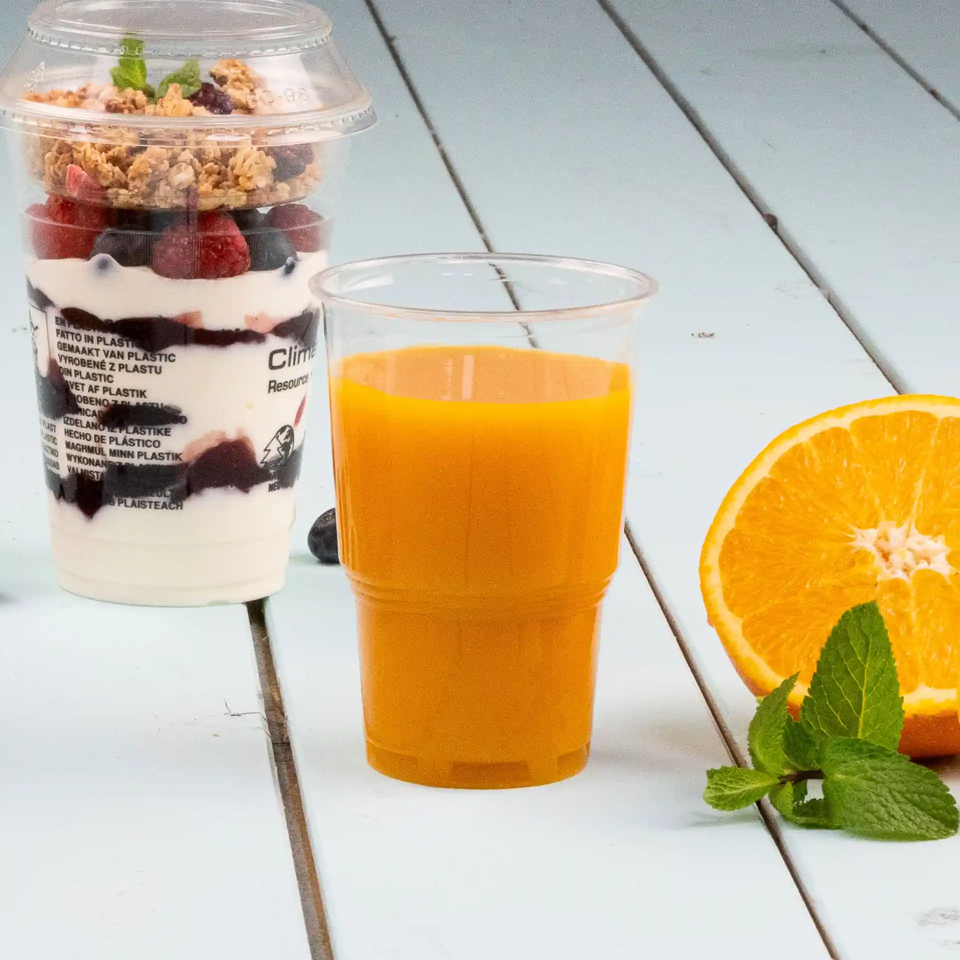 Image d'un gobelet en plastique transparent rempli de jus d'orange, à côté d'un parfait dans un gobelet avec granola et baies.