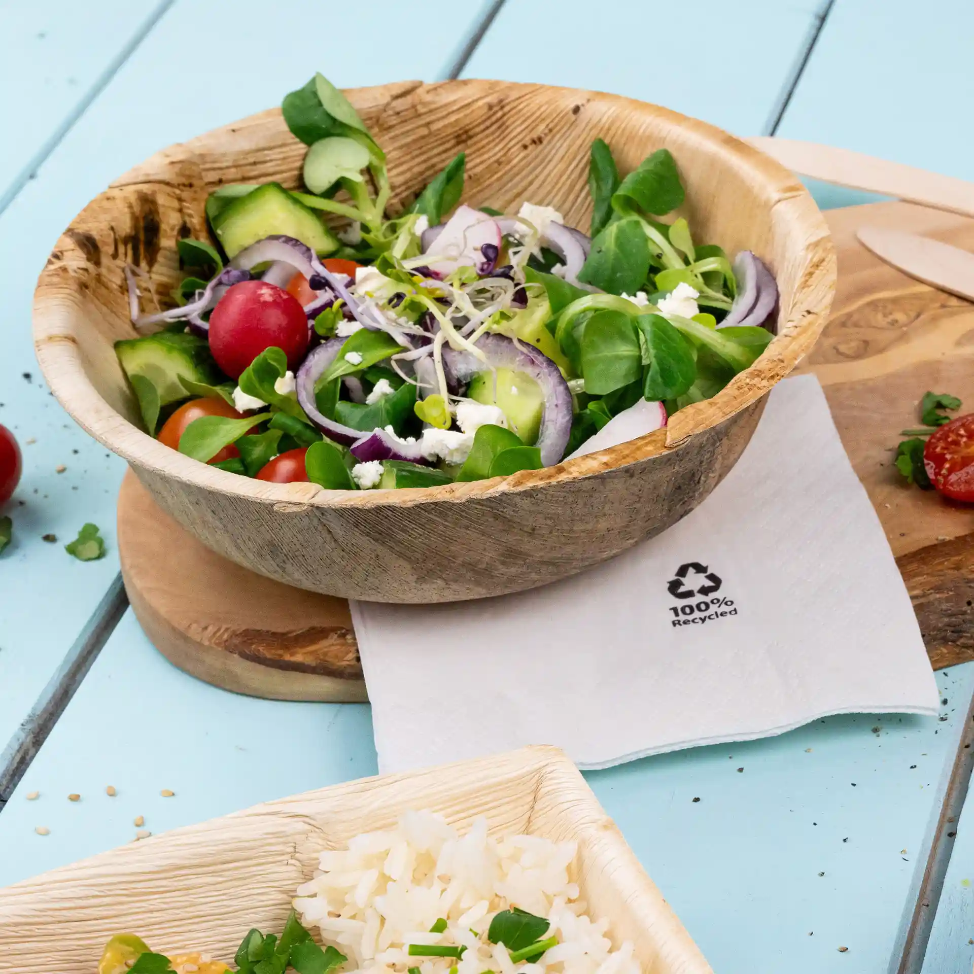 Bol rond en feuille de palmier rempli de salade mélangée, placé sur une planche en bois à côté d'une serviette et de riz dans un contenant.