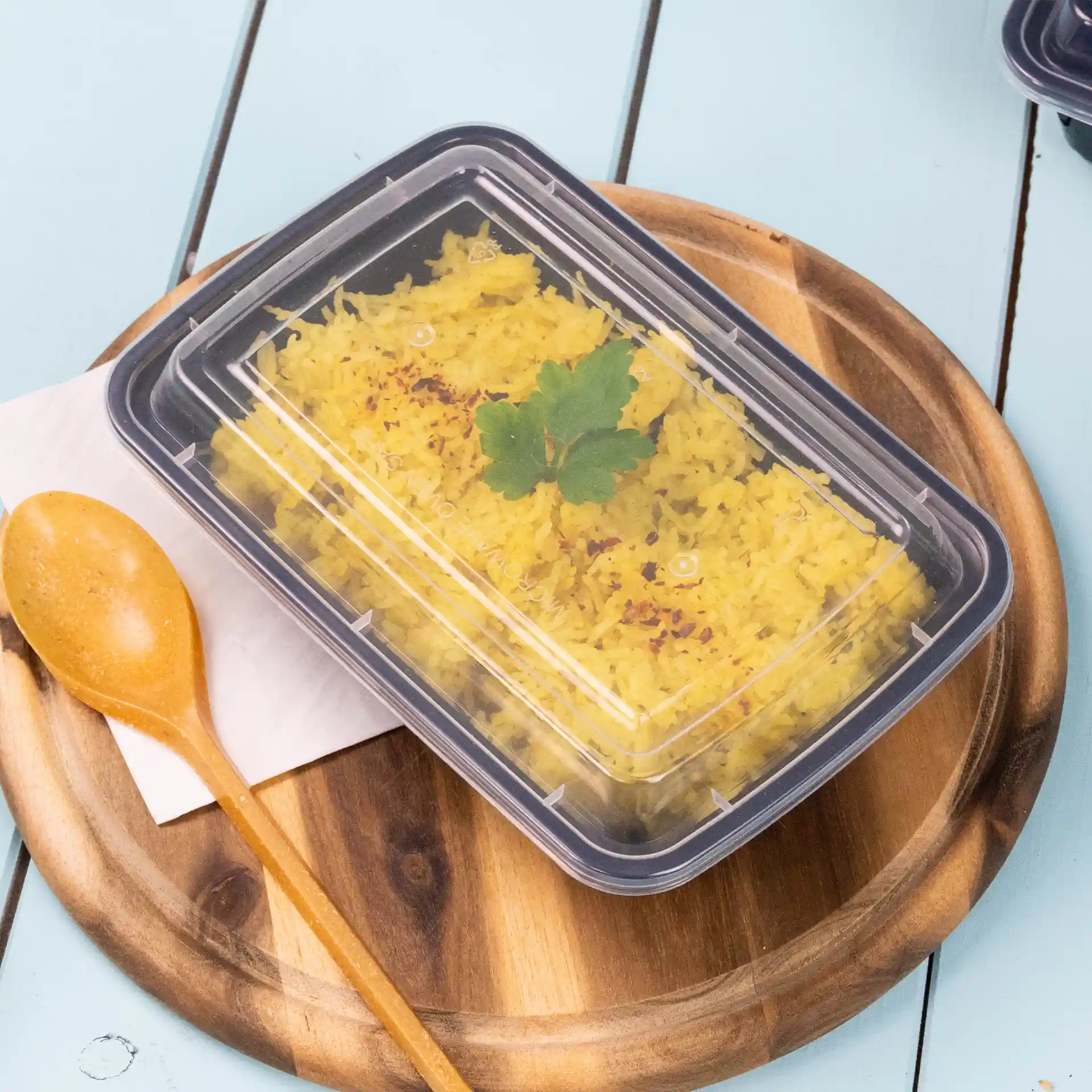 Conteneur rectangulaire transparent avec un couvercle réutilisable, contenant du riz jaune et garni d'une feuille de coriandre, sur une assiette en bois.