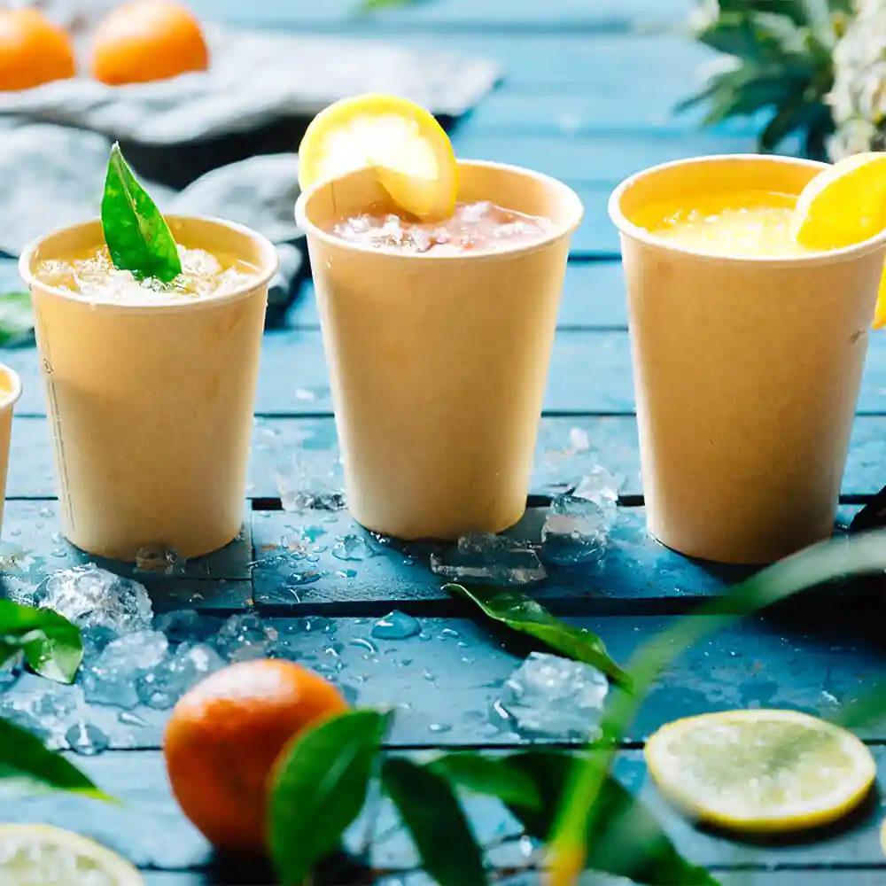 Trois gobelets en papier marron de 12 oz remplis de boissons, garnis de citron et de feuilles, sur une surface en bois bleu avec de la glace.