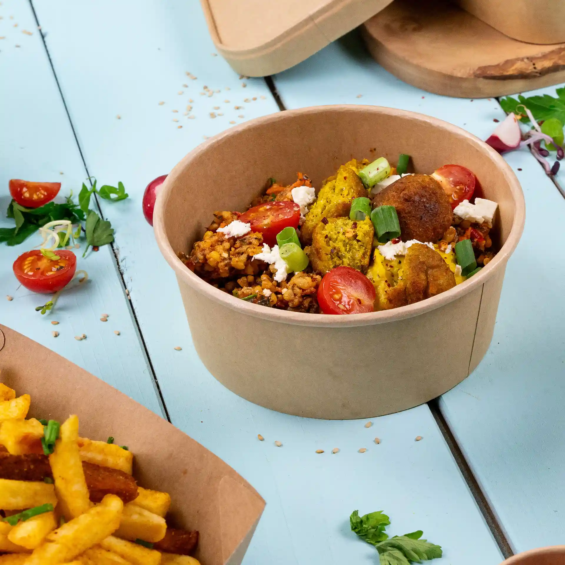 Bol en papier brun rond rempli de riz, légumes et tomates cerises, placé sur une surface bleue avec des frites à proximité.