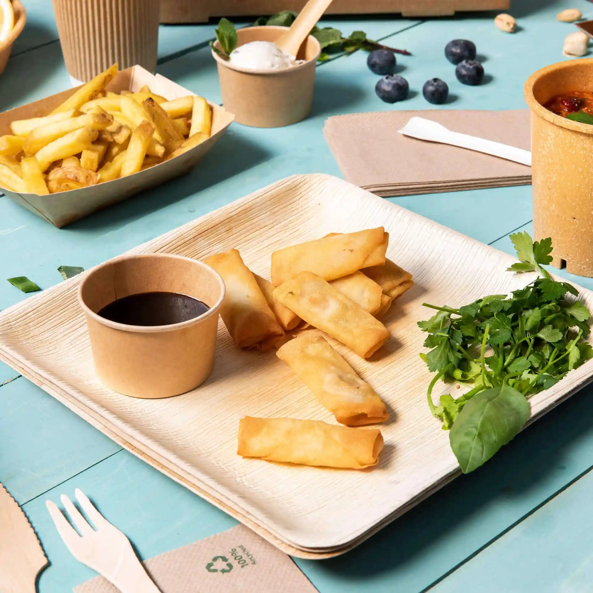 Une assiette carrée en feuille de palmier avec des rouleaux de printemps, un bol de sauce, des herbes fraîches et des frites sur une table bleue.