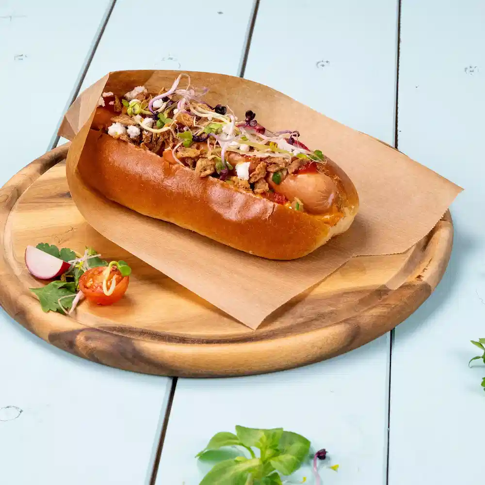 Hot-dog dans un pain garni d'oignons, d'herbes et de garnitures croquantes sur un plateau en bois avec des légumes d'accompagnement.