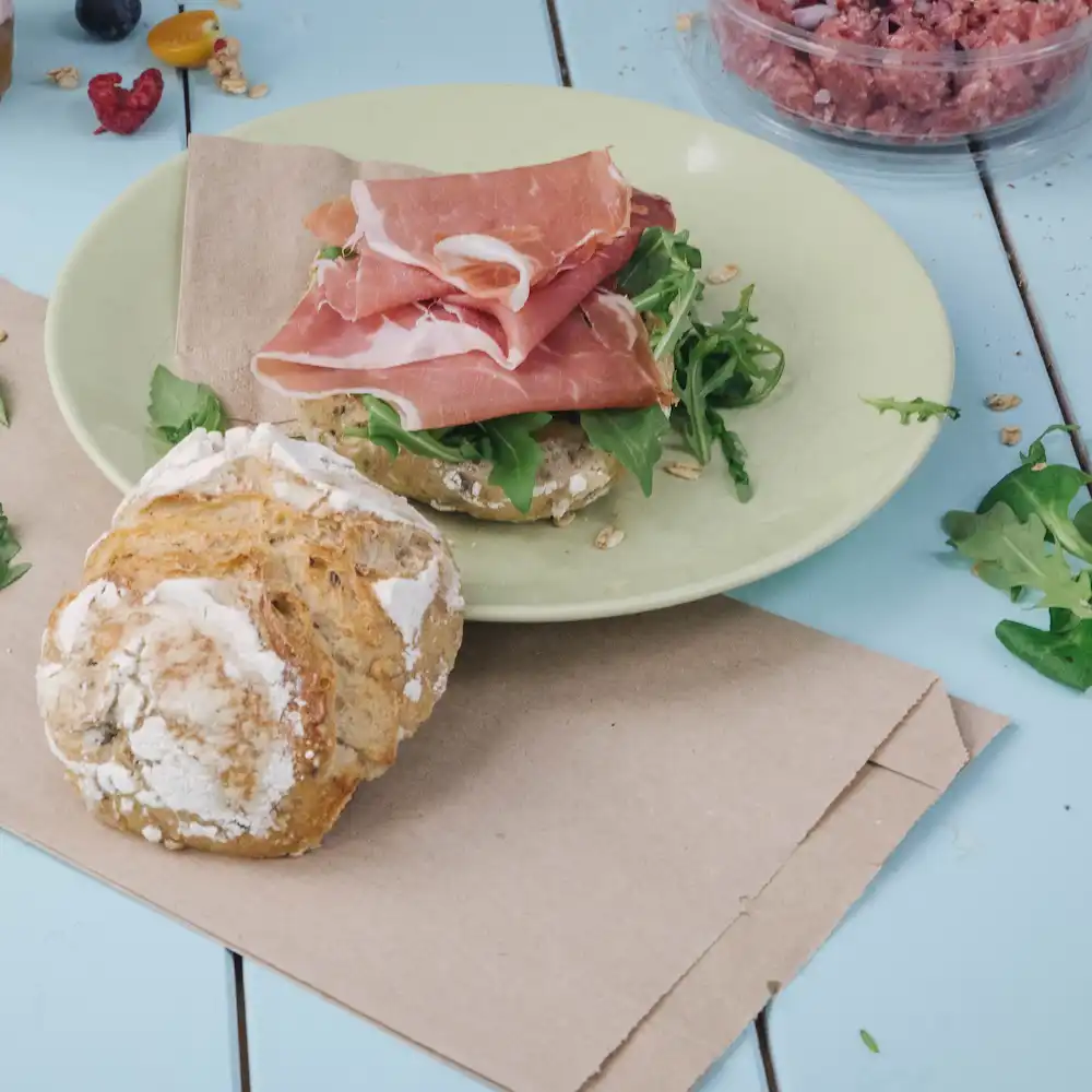 Tranches de prosciutto sur une assiette verte avec de la roquette sur un petit pain brun ; un petit rouleau de pain rond est à proximité.