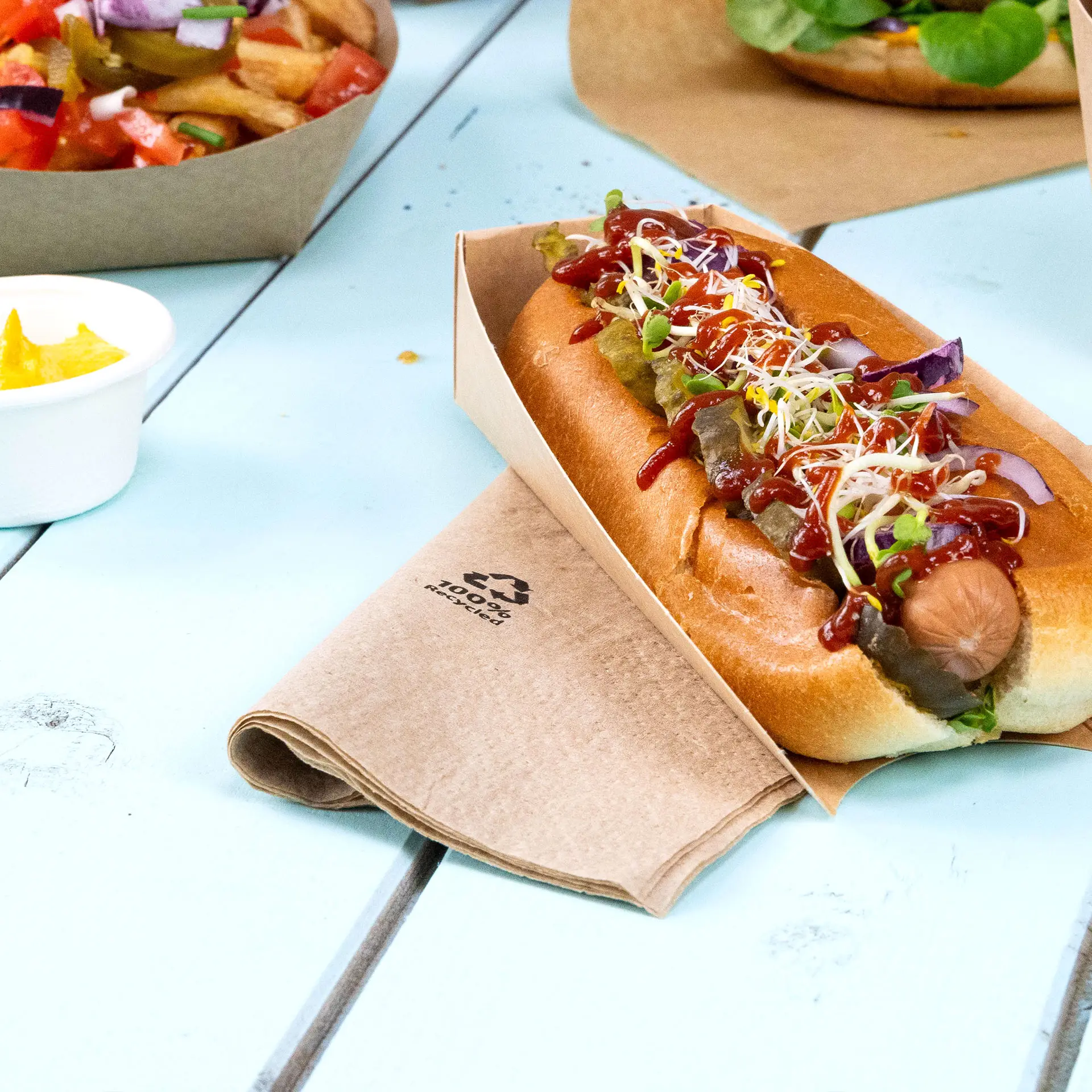 Un hot-dog dans un support en papier avec des garnitures, y compris du ketchup et des pousses ; serviette en papier recyclé visible à côté.