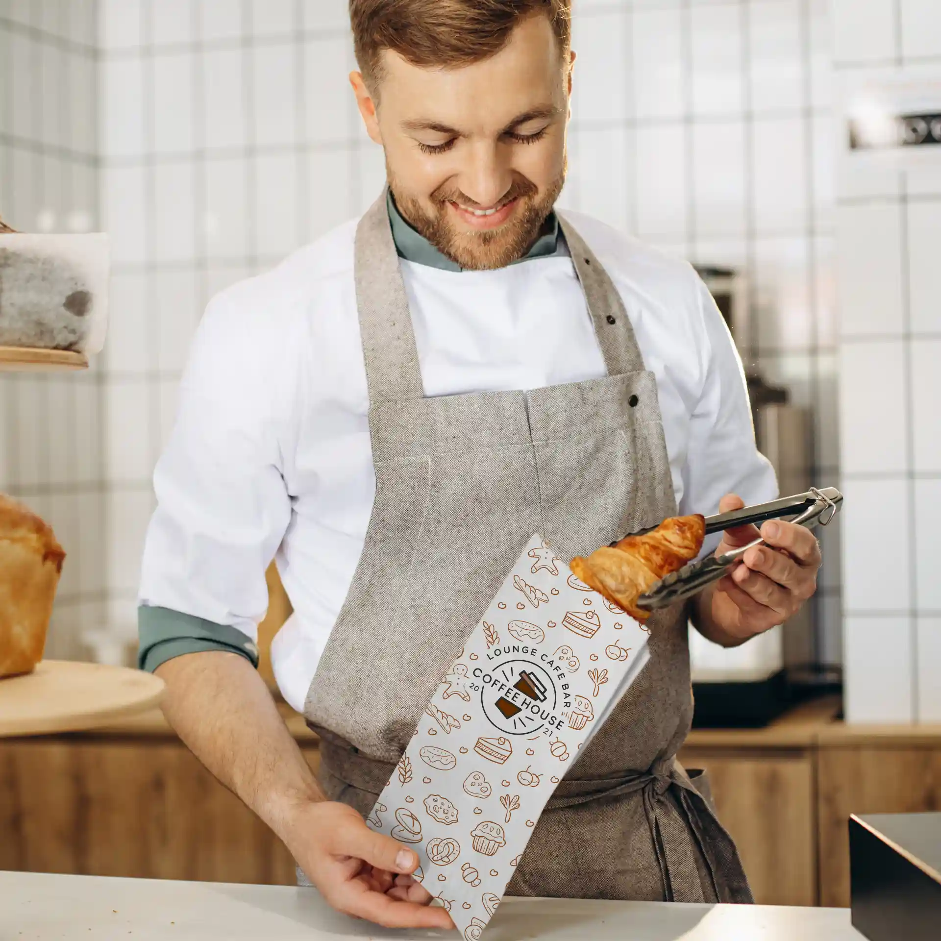 Image d'un homme en tablier tenant un sac en papier imprimé avec un croissant, dans un café.
