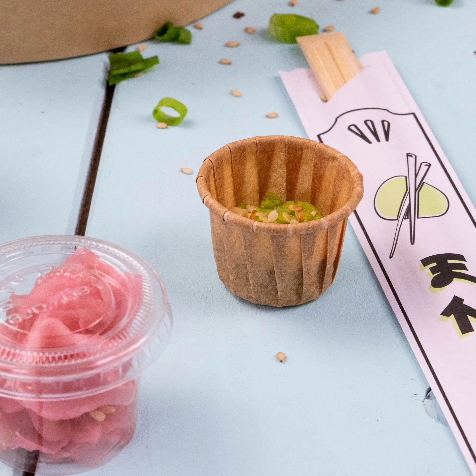 Gobelet en papier brun avec garniture verte et graines de sésame, accompagné de gingembre mariné et d'un étui à baguettes sur une surface bleue.