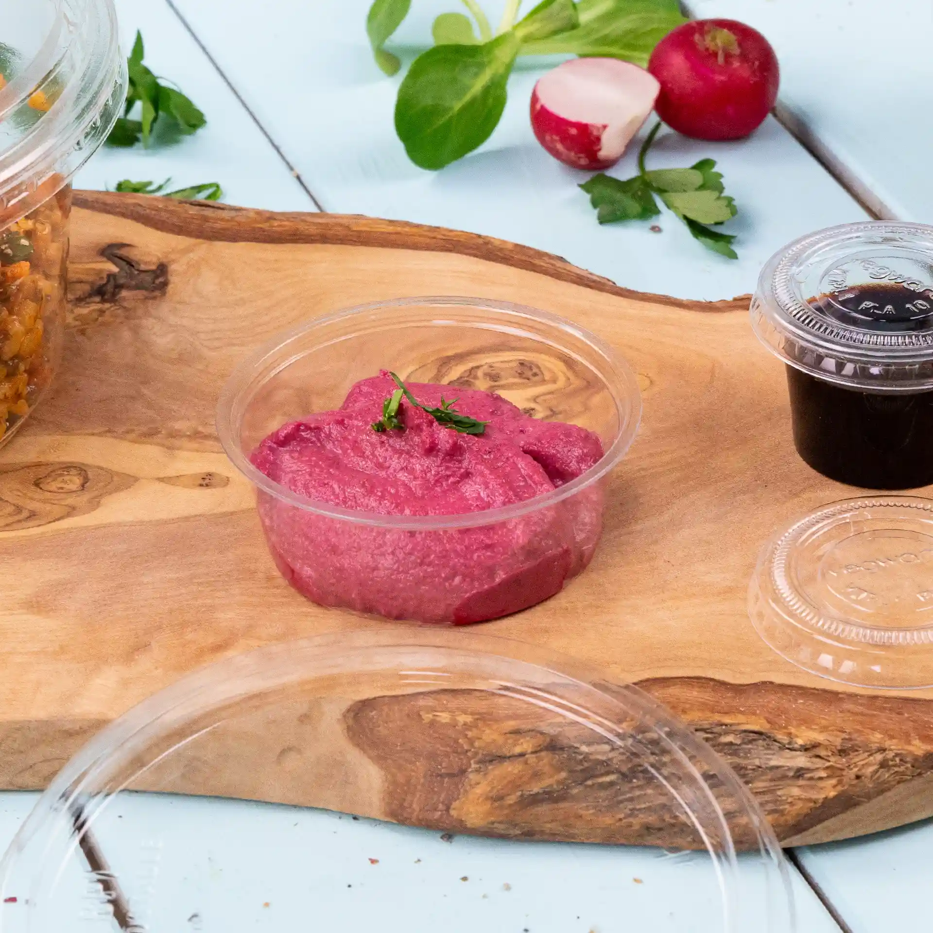 Pot à dip rond transparent avec un dip rose, garni d'une petite herbe verte, sur une surface en bois avec des légumes.