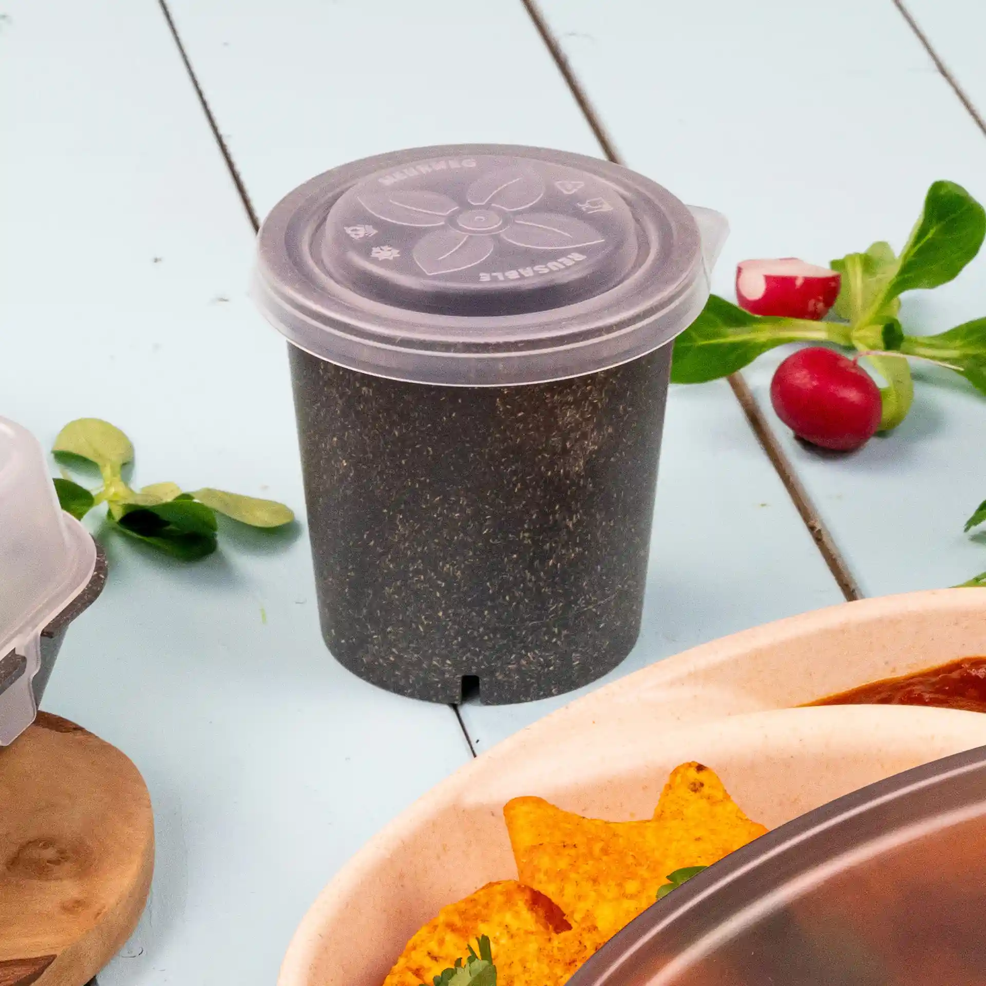 Gobelet rond transparent avec couvercle, en PP, posé sur une surface bleue avec des nachos et des légumes à proximité.