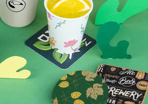 Des piles de sous-verres sur une surface en bois ; certains comportent un logo avec un oiseau et d'autres un design avec de la bière et du texte.