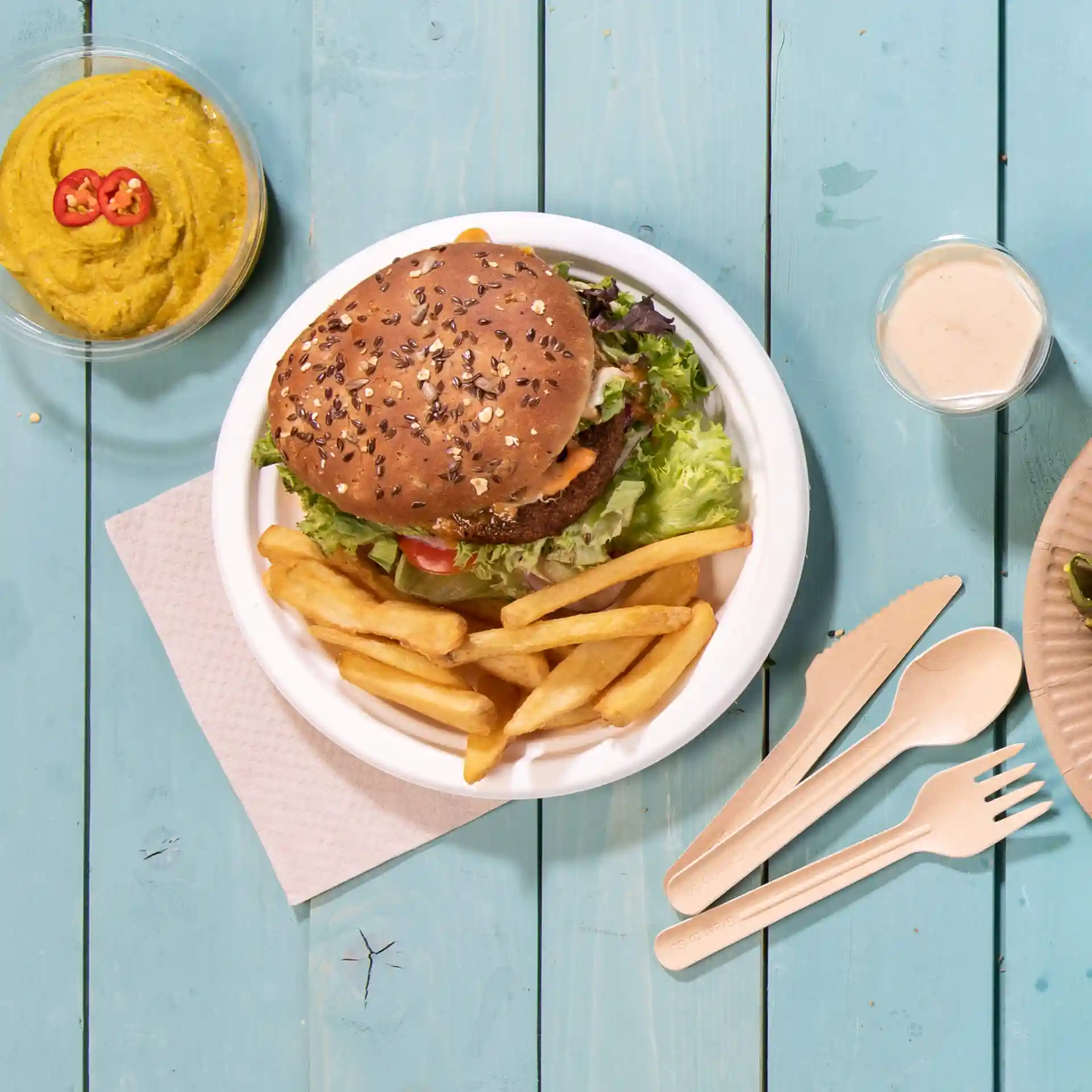 Image d'une assiette ronde en canne à sucre blanche avec un burger, des frites et un bol de houmous sur une surface en bois bleu.