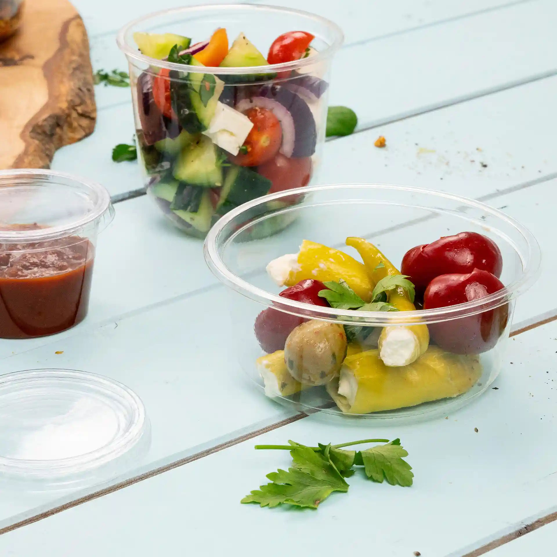 Conteneur rond transparent avec des légumes assortis, des olives et des herbes sur une surface en bois bleu.
