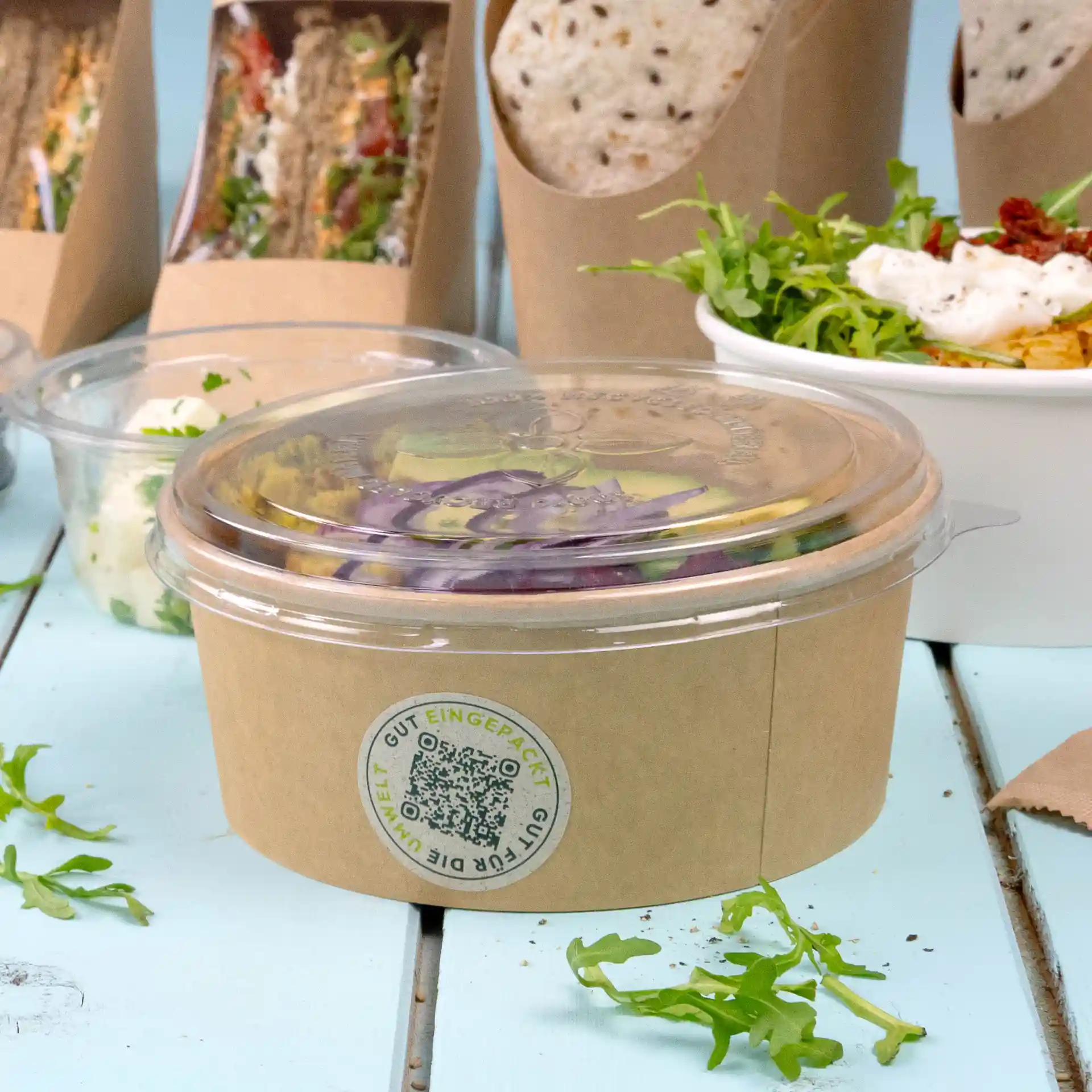 Gobelet rond transparent avec un couvercle transparent, contenant une salade mixte, posé sur une surface en bois bleu clair.