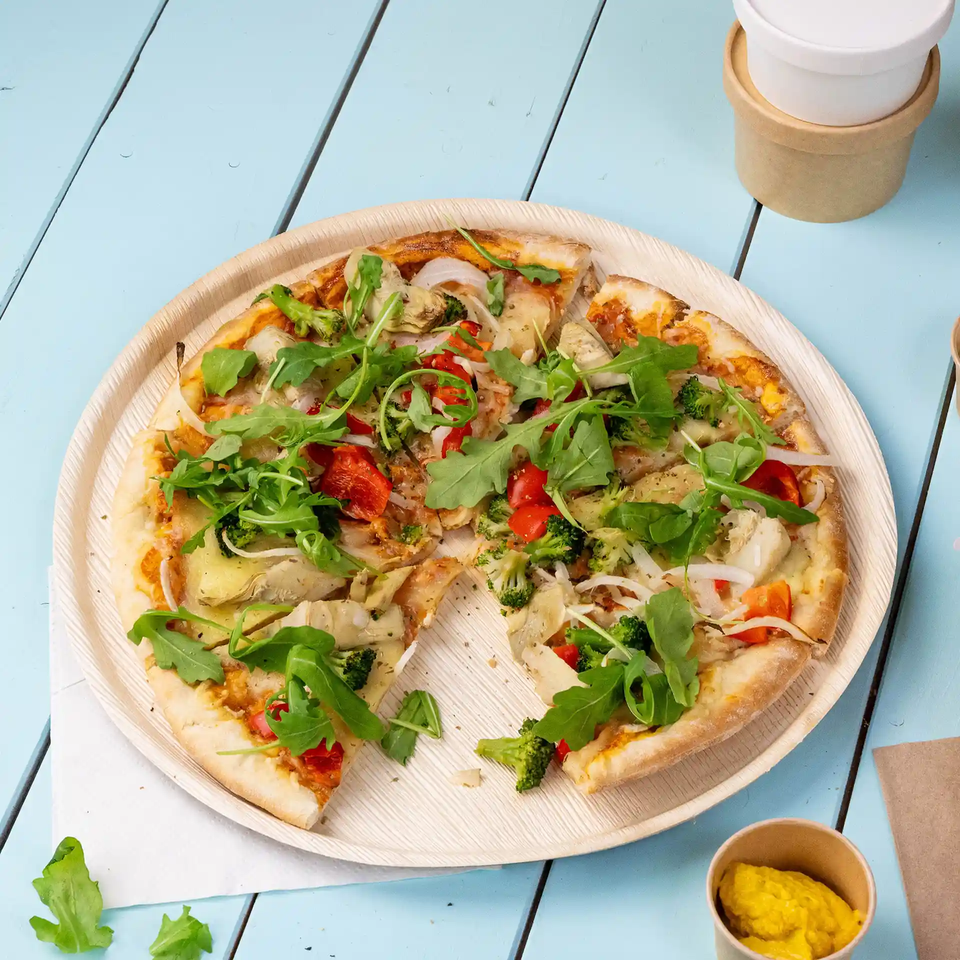 Assiette à pizza ronde en feuille de palmier de 27 cm de diamètre, contenant une pizza tranchée garnie de légumes et de roquette.