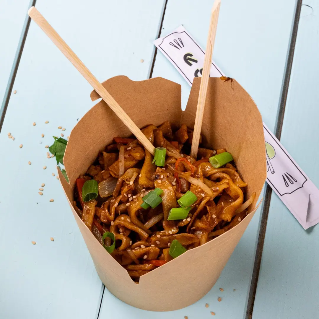 Conteneur en papier brun rempli de nouilles sautées, garni d'oignons verts et de graines de sésame, avec des baguettes insérées.