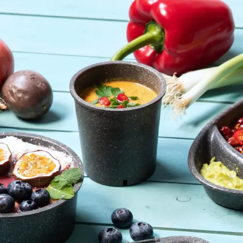 Tasse à soupe réutilisable remplie de soupe et garnie d'herbes, entourée de légumes et de fruits frais sur une surface bleue.