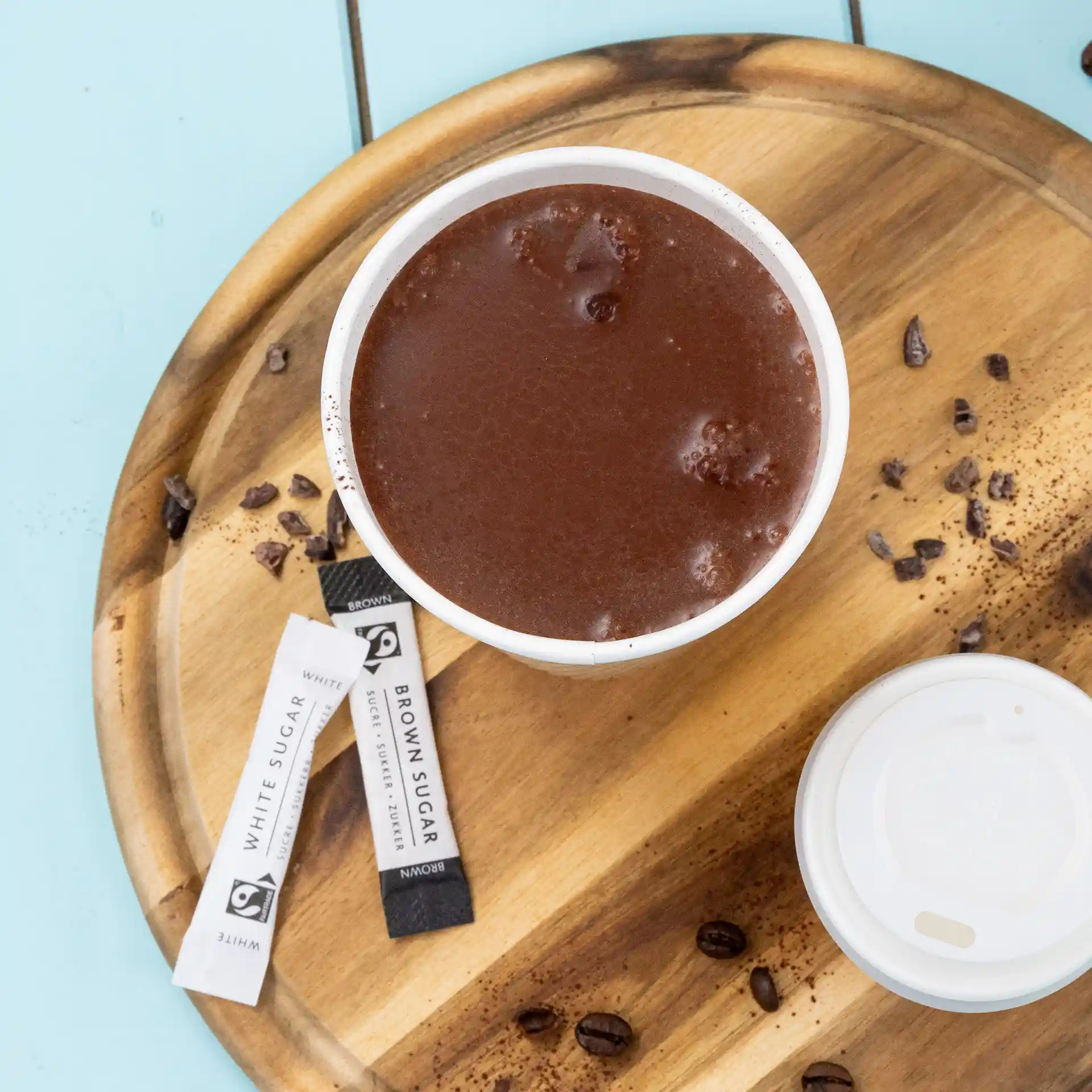 Tasse de chocolat chaud sur un plateau en bois avec des sachets de sucre et des grains de café éparpillés.
