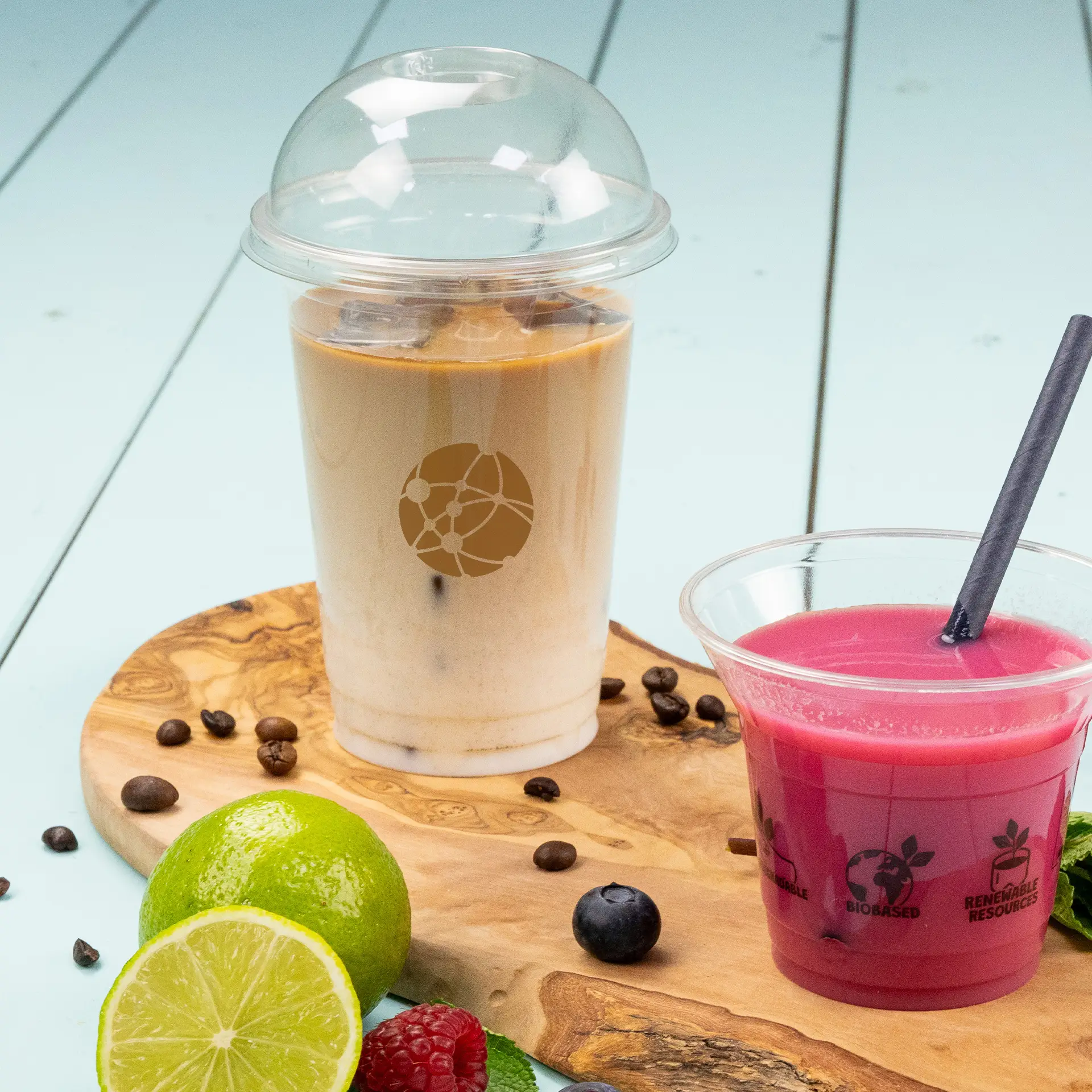 Un gobelet PLA transparent rempli de café glacé à côté d'un smoothie rose; citron vert frais, baies et grains de café sur une planche en bois.