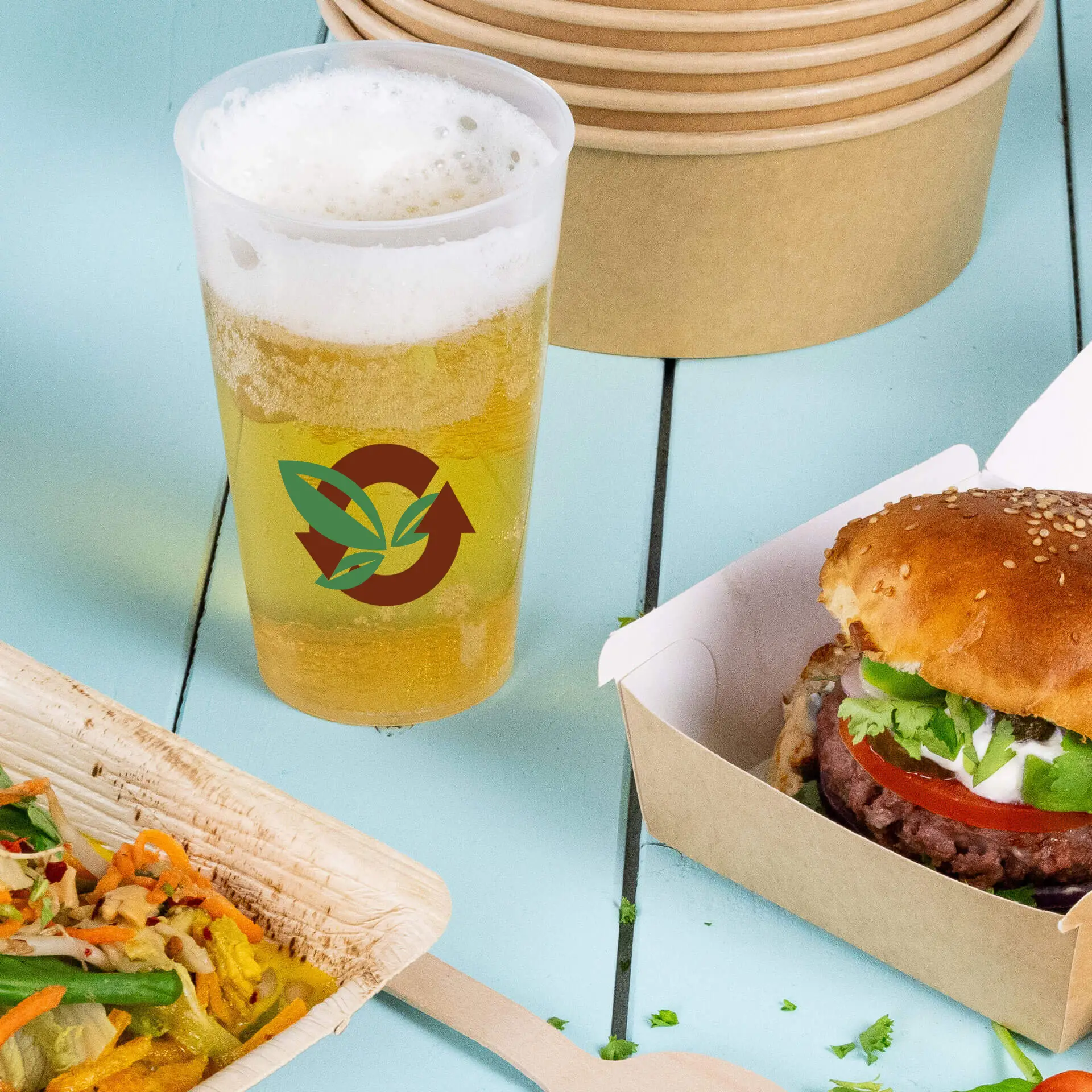 Gobelet réutilisable de 400 ml avec logo et bière mousseuse, à côté d'un burger et d'une salade sur une table en bois bleue.