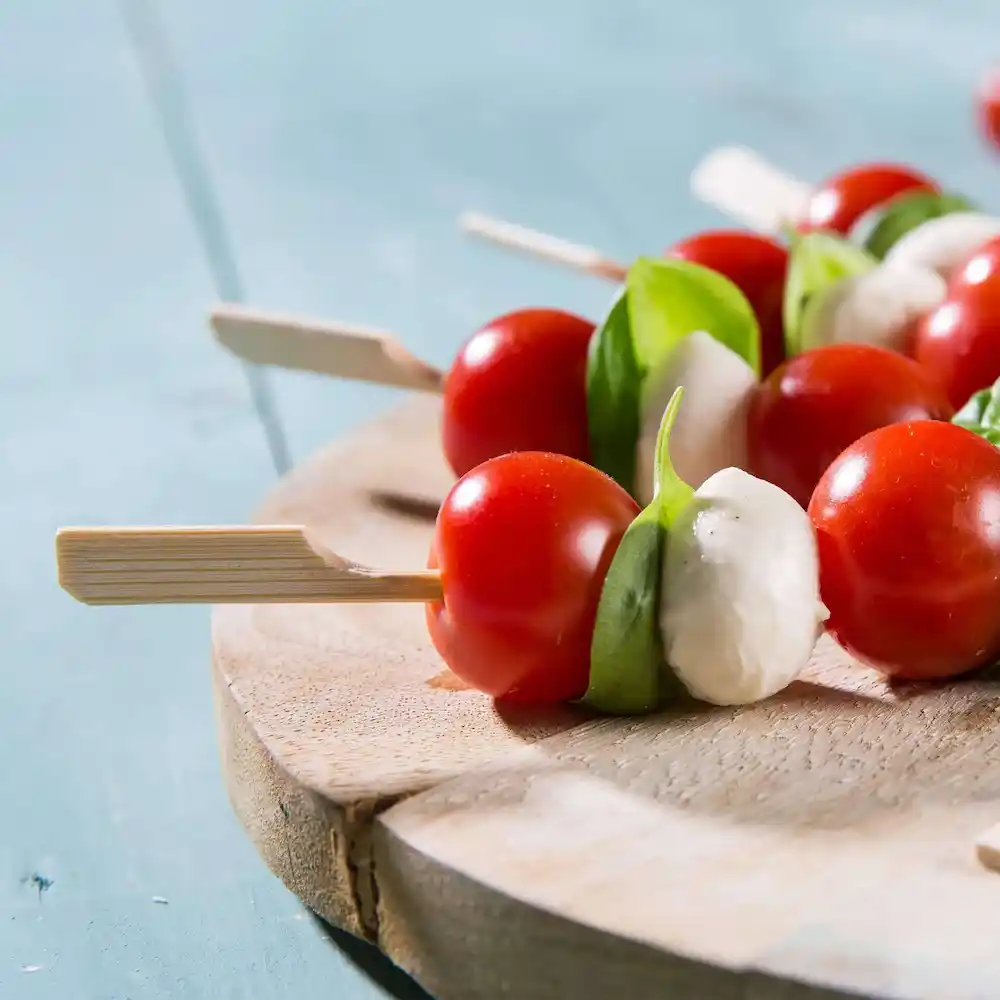 Brochettes de tomates cerises, feuilles de basilic et boules de mozzarella disposées sur une planche de service en bois.