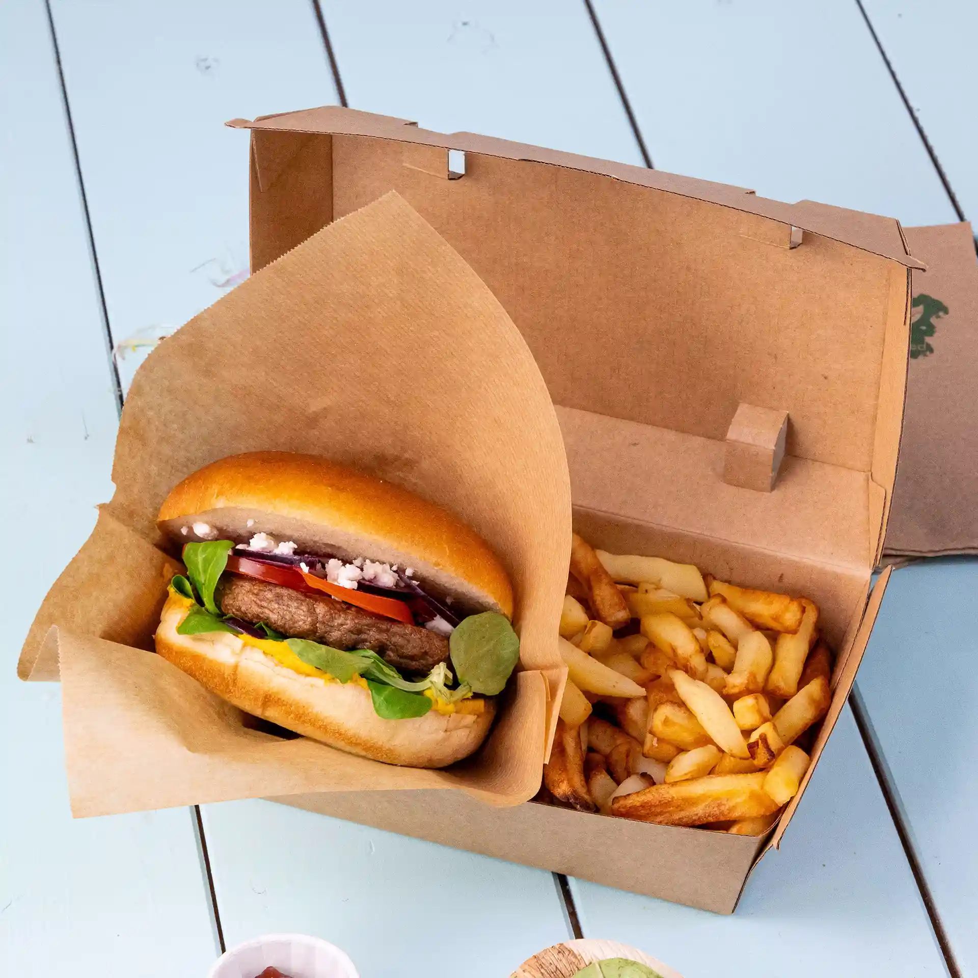 Boîte en carton marron contenant un hamburger avec garnitures et des frites, sur une surface bleu clair.