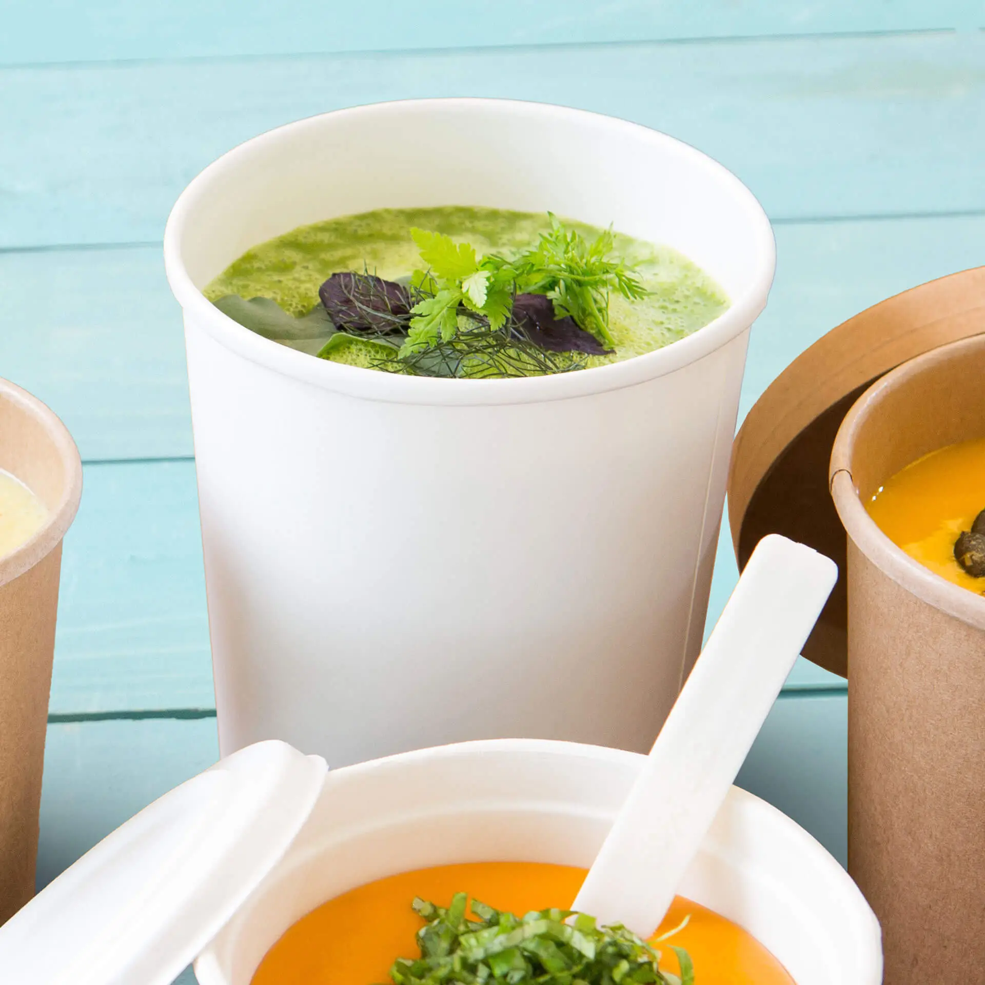 Gobelet à soupe biodégradable blanc avec soupe verte et herbes, placé avec d'autres contenants de soupe sur un fond bleu sarcelle.