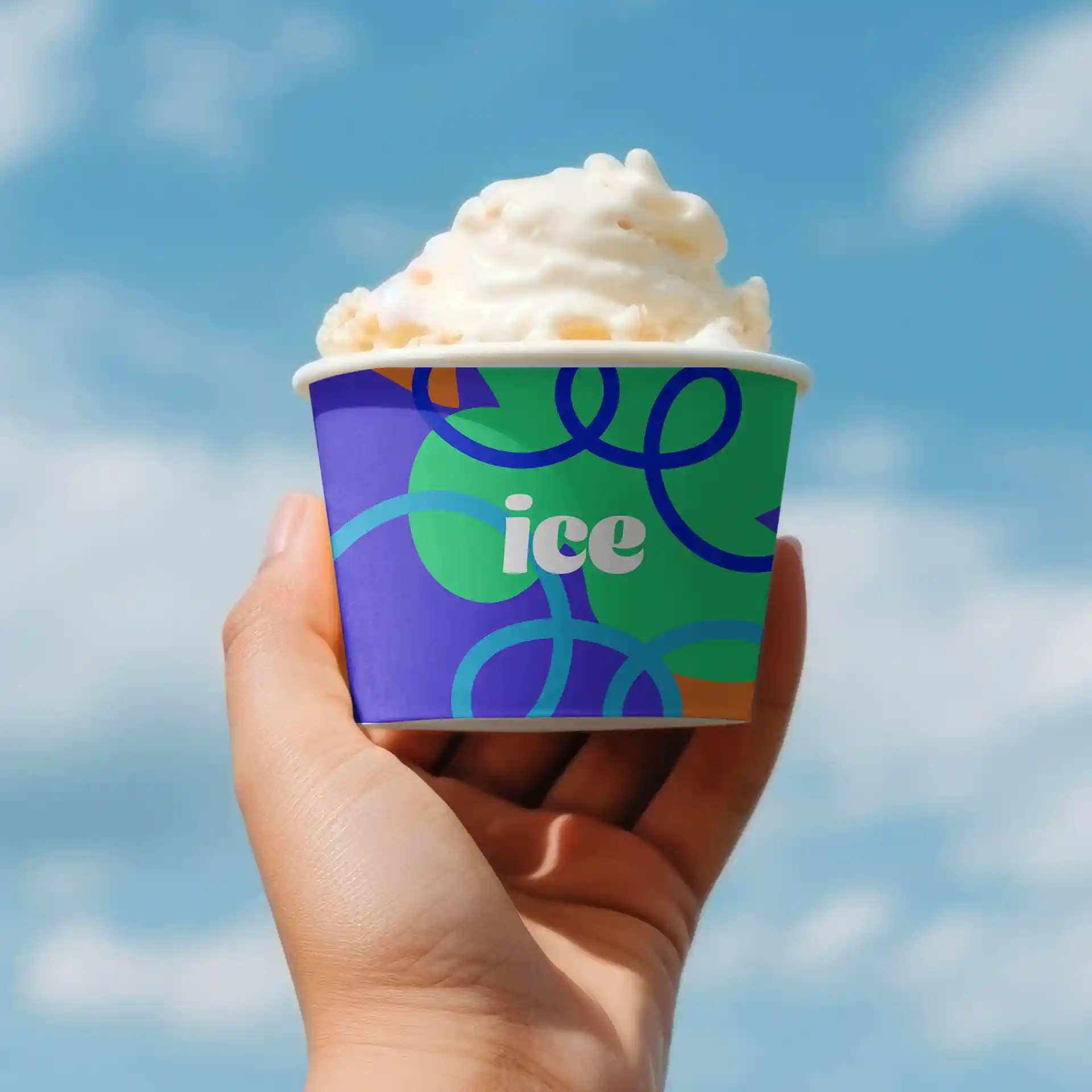 Gobelet en carton rond pour glace avec un design coloré, tenu dans une main contre un ciel bleu, rempli de glace.