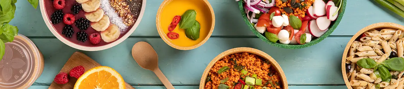 Des bols colorés remplis de divers aliments, y compris des fruits, des pâtes, des salades et des céréales, disposés sur une table en bois.