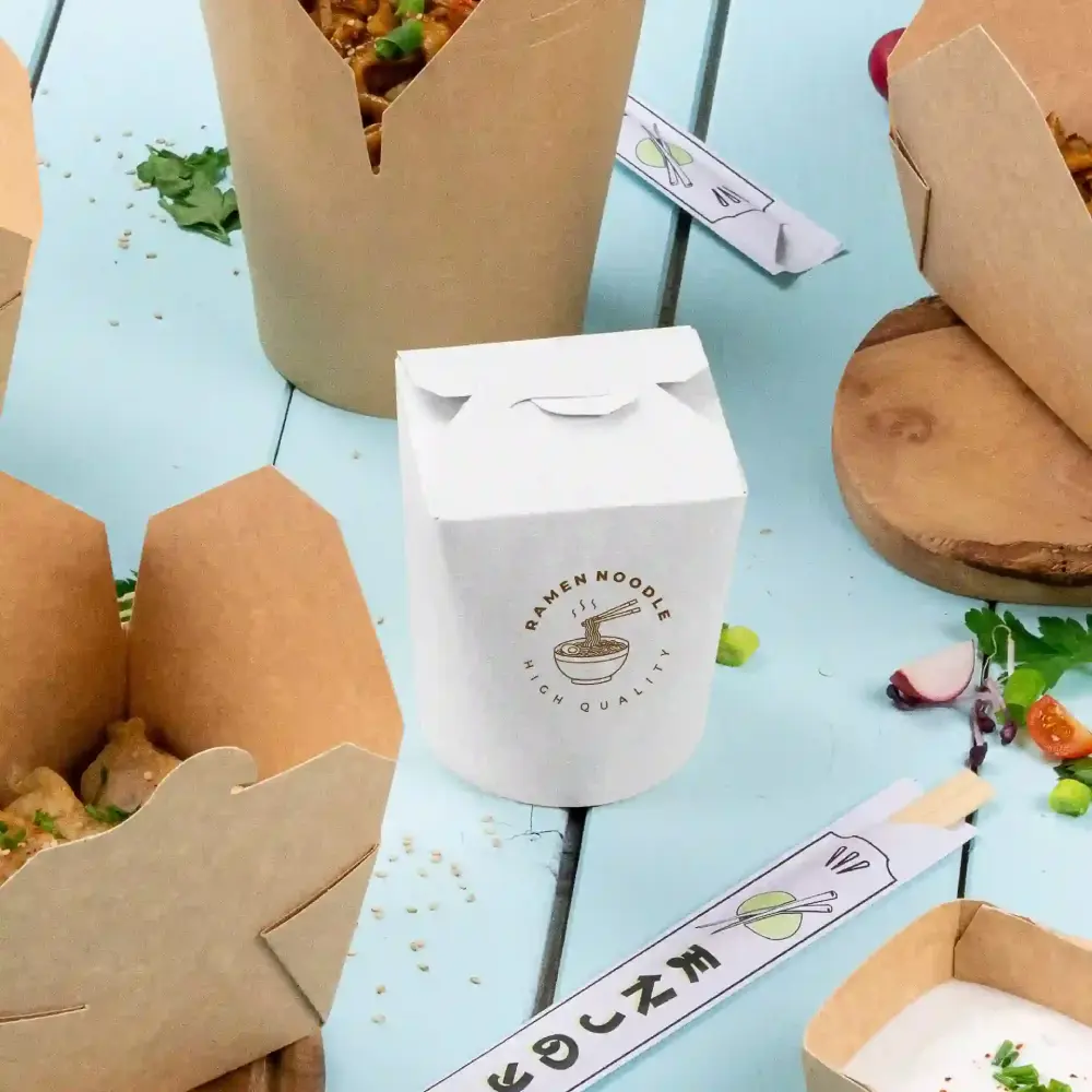 Boîte à emporter blanche étiquetée "Nouilles Ramen de haute qualité" à côté de boîtes à emporter brunes sur une surface bleue.