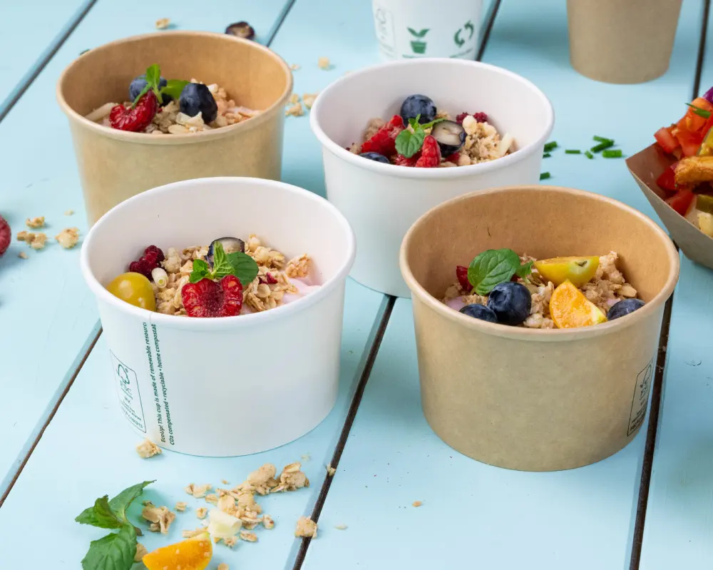 Quatre contenants alimentaires remplis de granola et de fruits frais, y compris des baies et des agrumes, sur une surface en bois bleue.