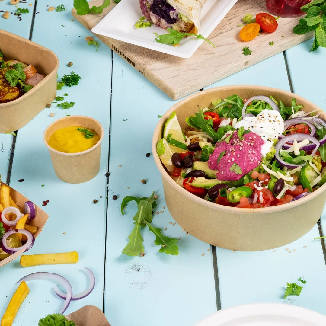 Bol de salade avec légumes mélangés, citron vert et sauces, entouré de petits contenants de sauce, frites et herbes sur une surface bleue.