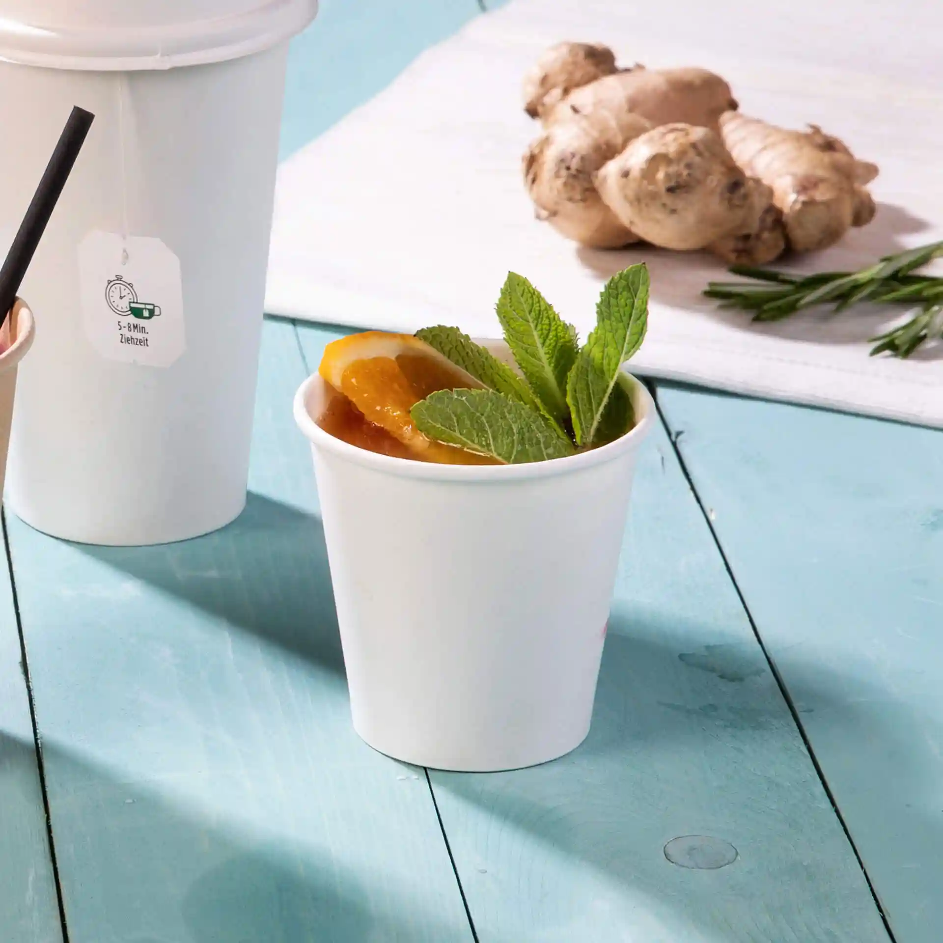 Gobelet blanc rempli d'une boisson, garni de tranches d'orange et de feuilles de menthe, posé sur une surface en bois bleu.