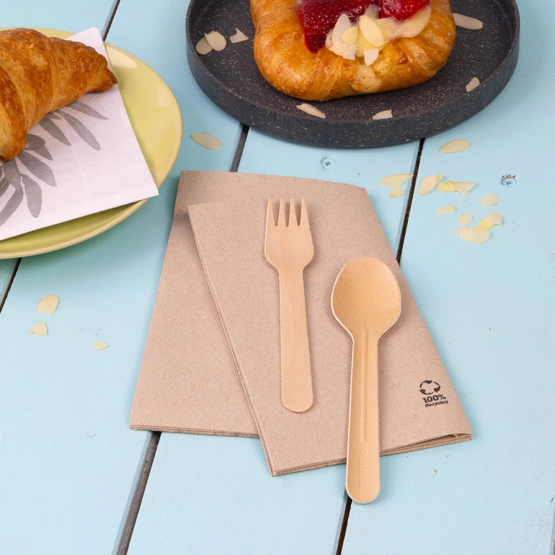Serviettes en papier recyclé marron pliées avec une cuillère et une fourchette biodégradables sur une surface bleue près d'un croissant et d'une pâtisserie.