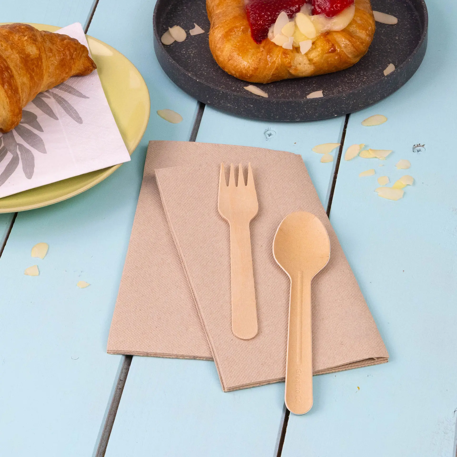 Deux serviettes en papier recyclé non blanchi avec une fourchette et une cuillère en bois sur une table bleue.