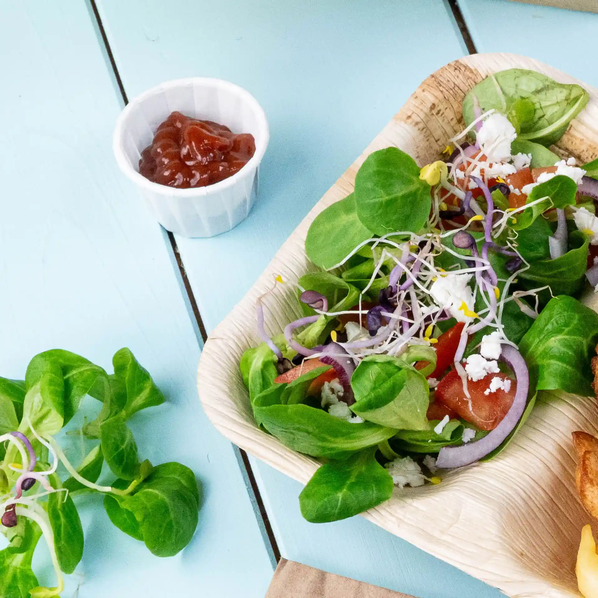 Salade avec des légumes mélangés, tomates, oignons et pousses dans un bol en feuille de palmier, accompagnée d'une petite tasse blanche de ketchup.