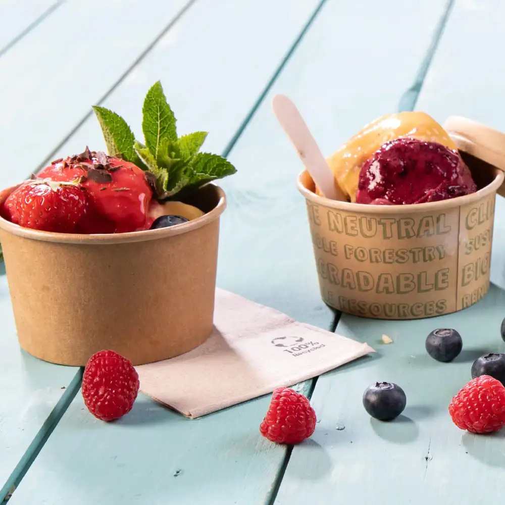 Deux coupes de glace avec des baies sur une surface en bois bleu, l'une garnie de fraises et de menthe, l'autre de glace violette et jaune.