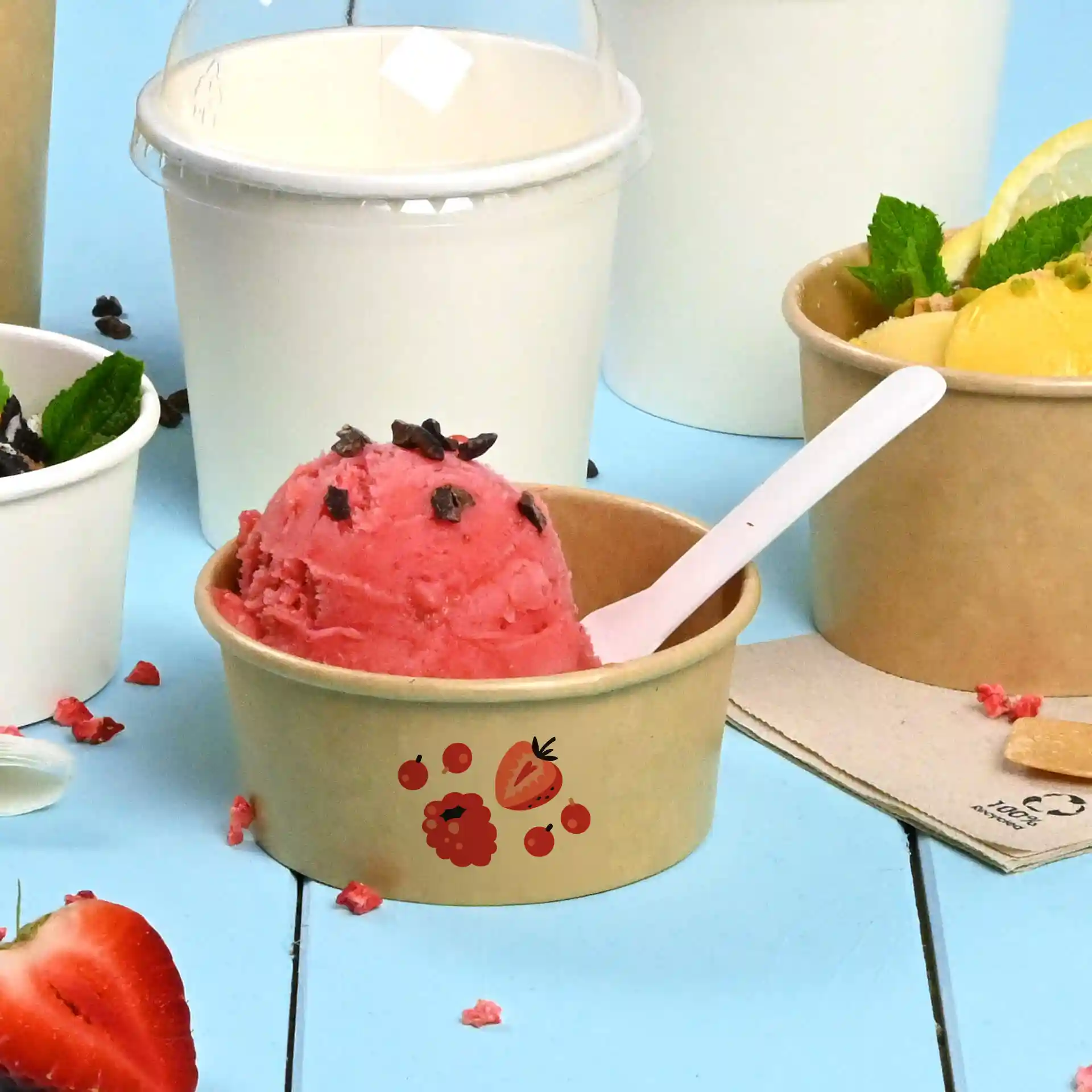 Gobelet en carton marron avec glace rose surmontée de morceaux de chocolat et une cuillère blanche, sur fond bleu.