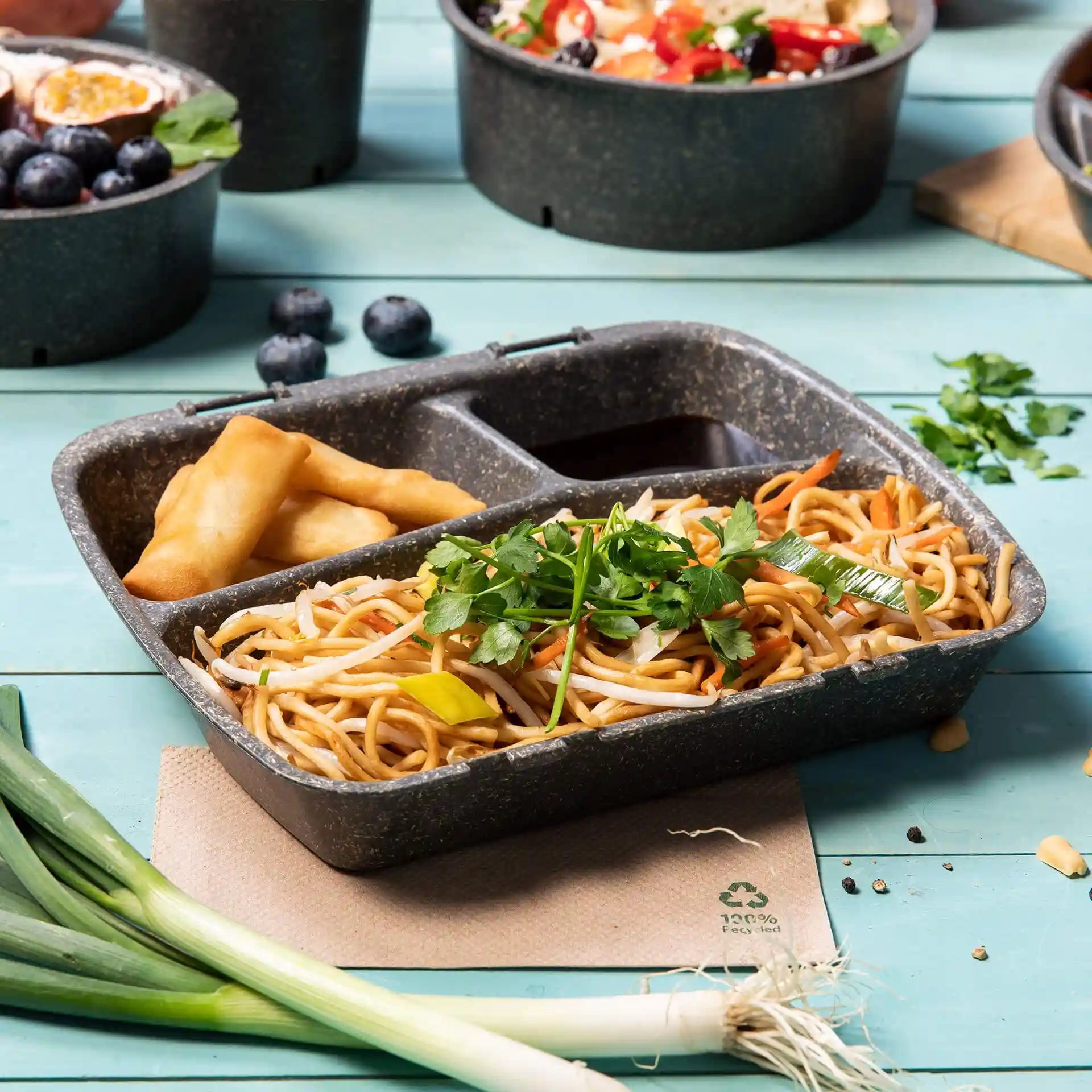 Conteneur de repas réutilisable gris avec trois compartiments, rempli de nouilles, de sauce et de rouleaux de printemps, sur une surface bleue.