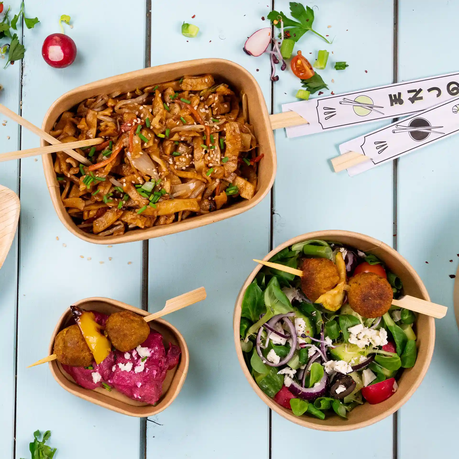 Image de trois contenants alimentaires : un grand avec des nouilles, un moyen avec de la salade et un petit avec du dip et des boulettes, le tout sur une surface bleue.