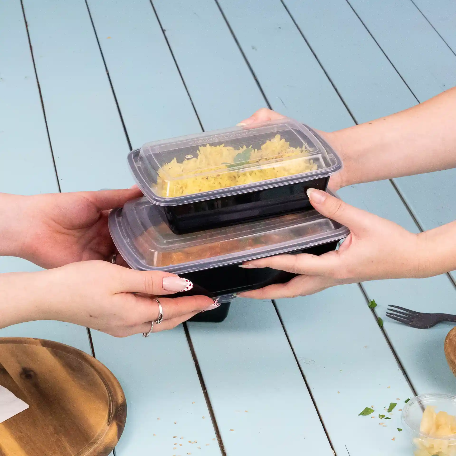 Deux mains tiennent des contenants alimentaires rectangulaires empilés avec des couvercles transparents, placés sur une surface bleu clair.