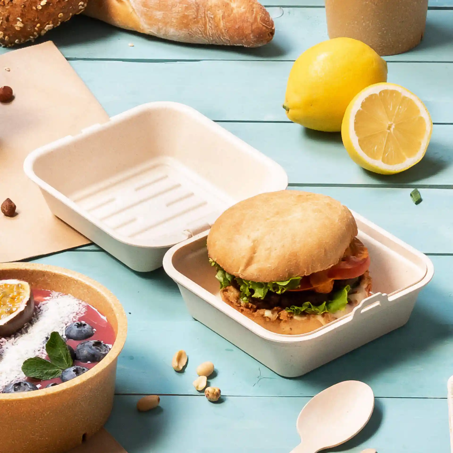 Boîte à burger réutilisable blanche crémeuse avec un sandwich, entourée d'un bol de fruits et d'autres aliments.